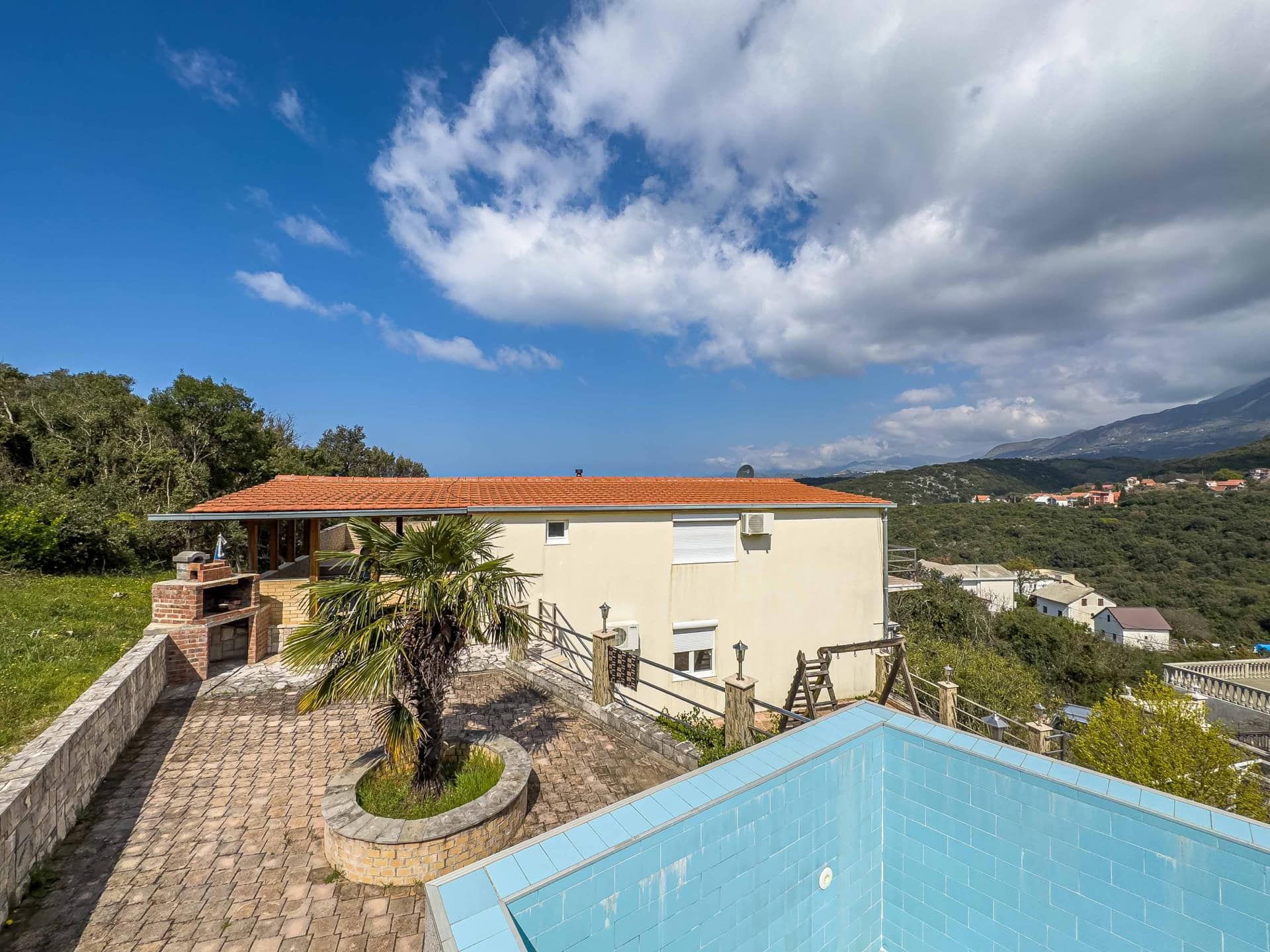 Two-story house in Utjeha, Montenegro: terrace with a pool, brick barbecue, palm tree, and panoramic mountain view.