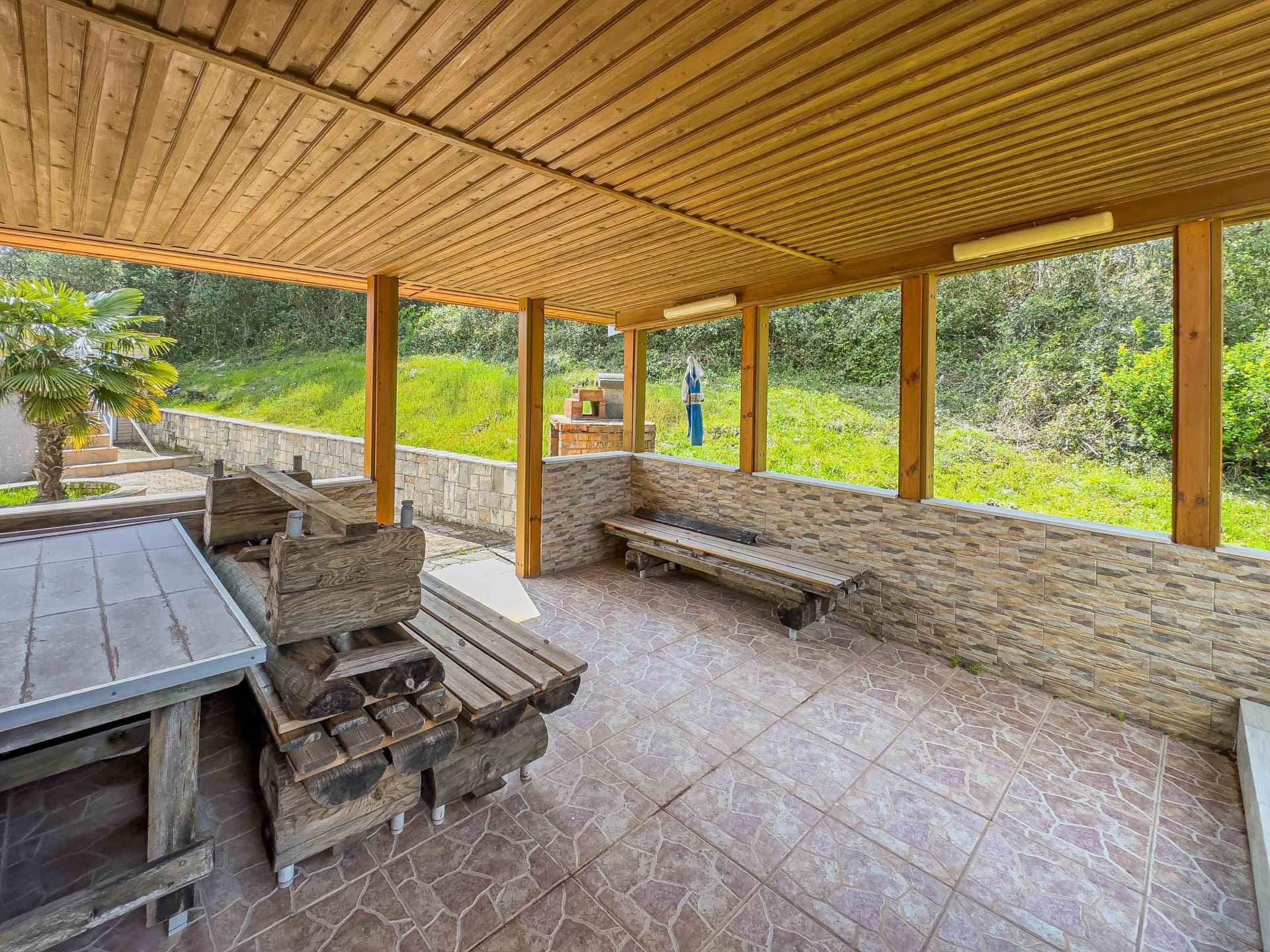 Covered terrace with wooden furniture and barbecue area in a house, Uteha, Montenegro, overlooking a green garden.