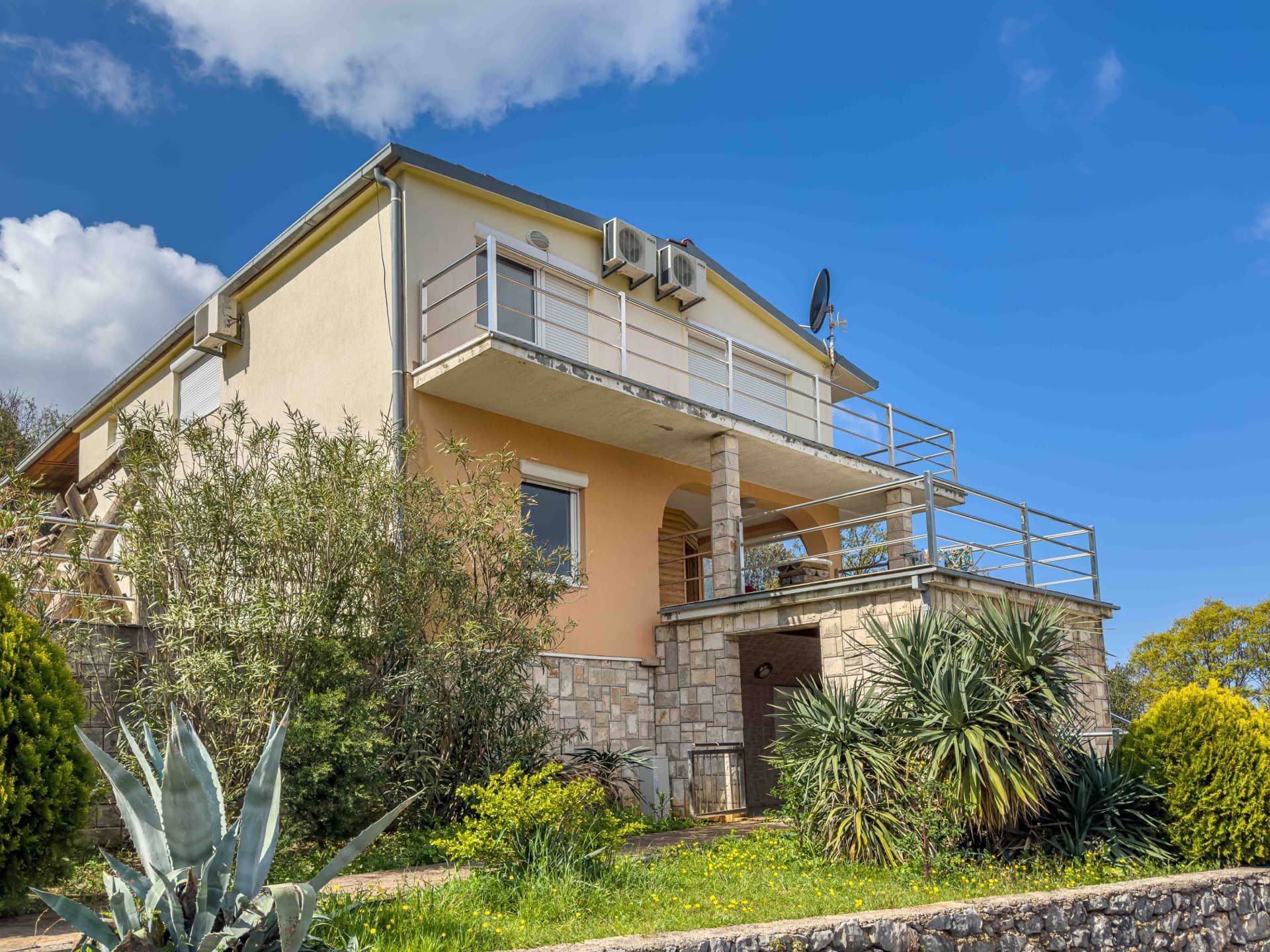 Two-story house in Utjeha, Montenegro: stone-clad facade, spacious balconies, and a garden against a blue sky.