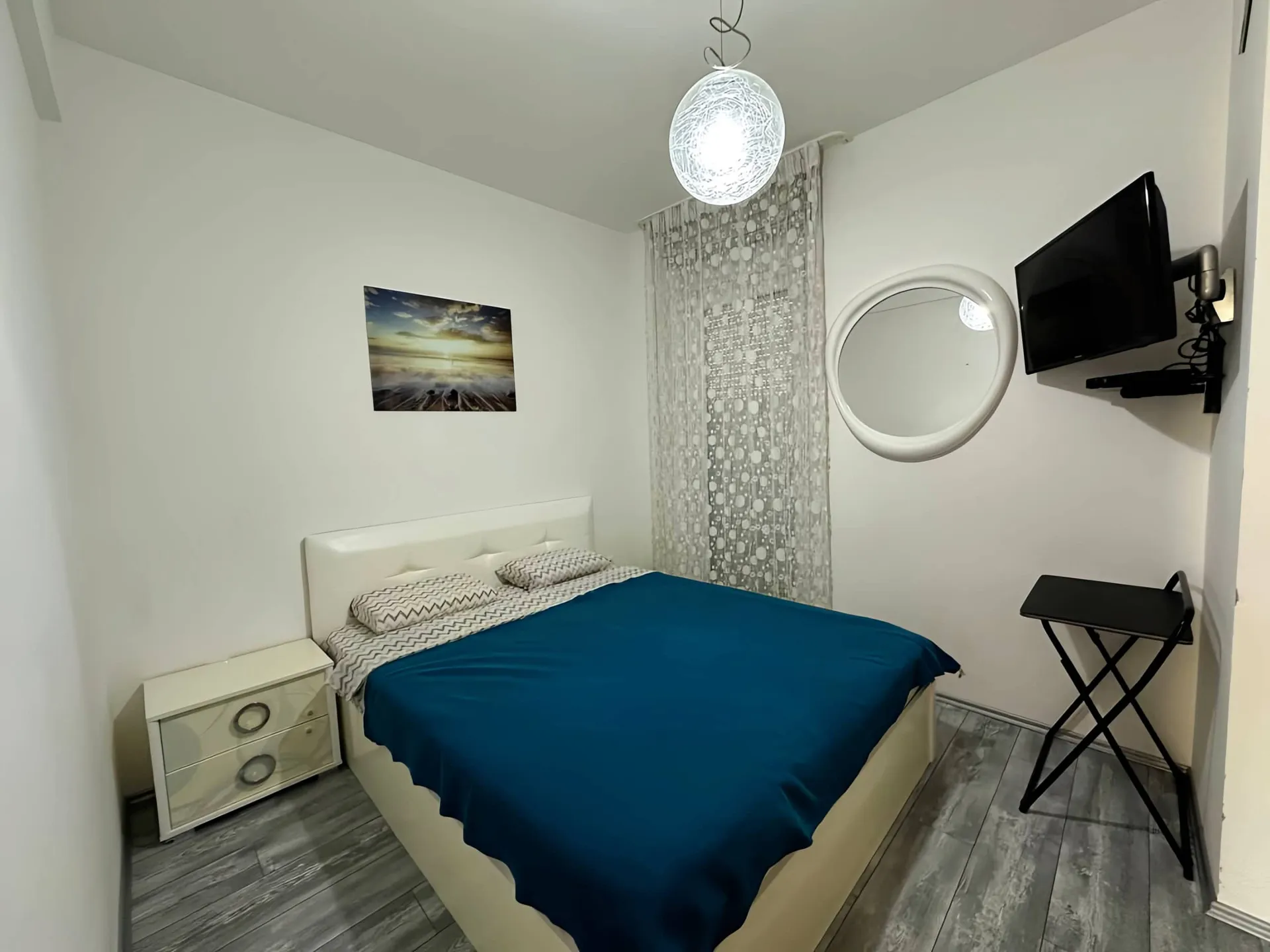 Bedroom featuring a double bed with blue bedding, white nightstand, wall-mounted TV, round mirror, and patterned curtains.