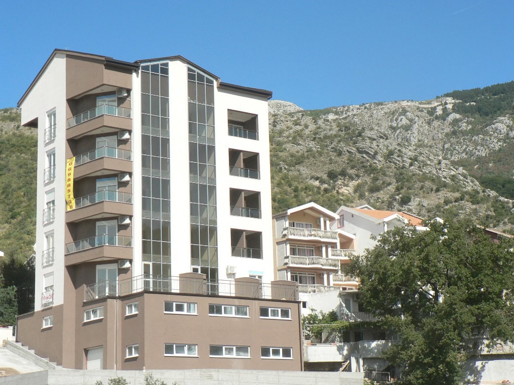 Modern residential building with panoramic windows and balconies in Bečići, Montenegro, against a mountain backdrop.
