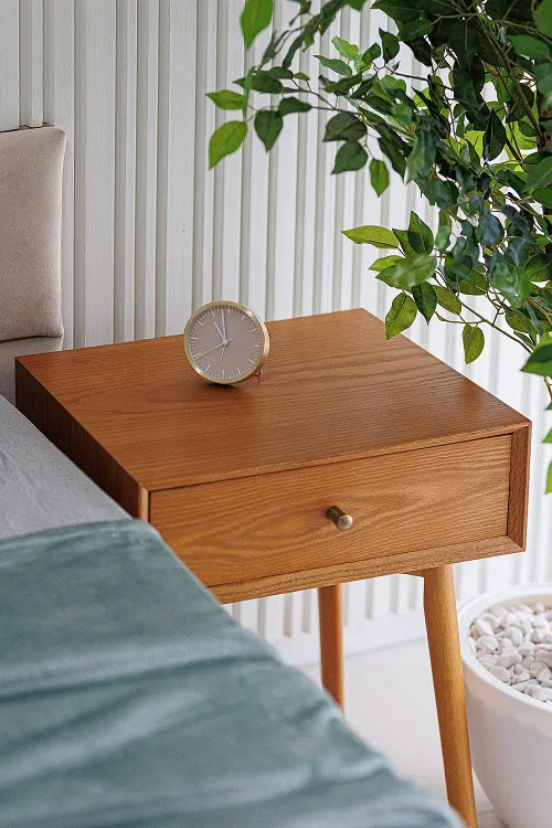 Wooden nightstand with a clock, part of a bed with bedding, and a green plant against a white wall.