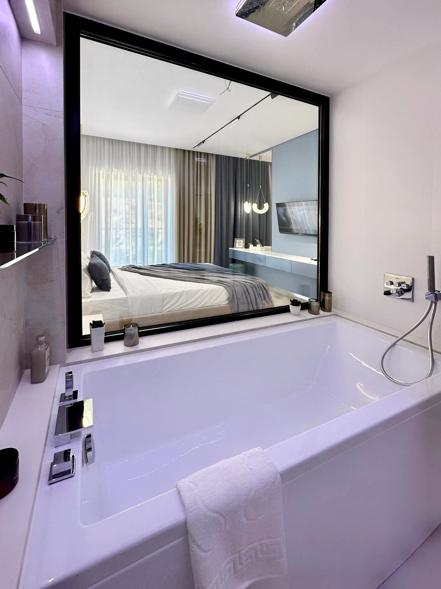White bathtub with chrome fixtures; a large window reflects an adjacent bedroom featuring a bed and curtains.