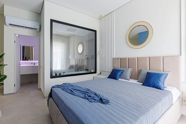 Modern bedroom featuring a large bed with blue bedding, a wall mirror, and an adjacent bathroom visible through a doorway.