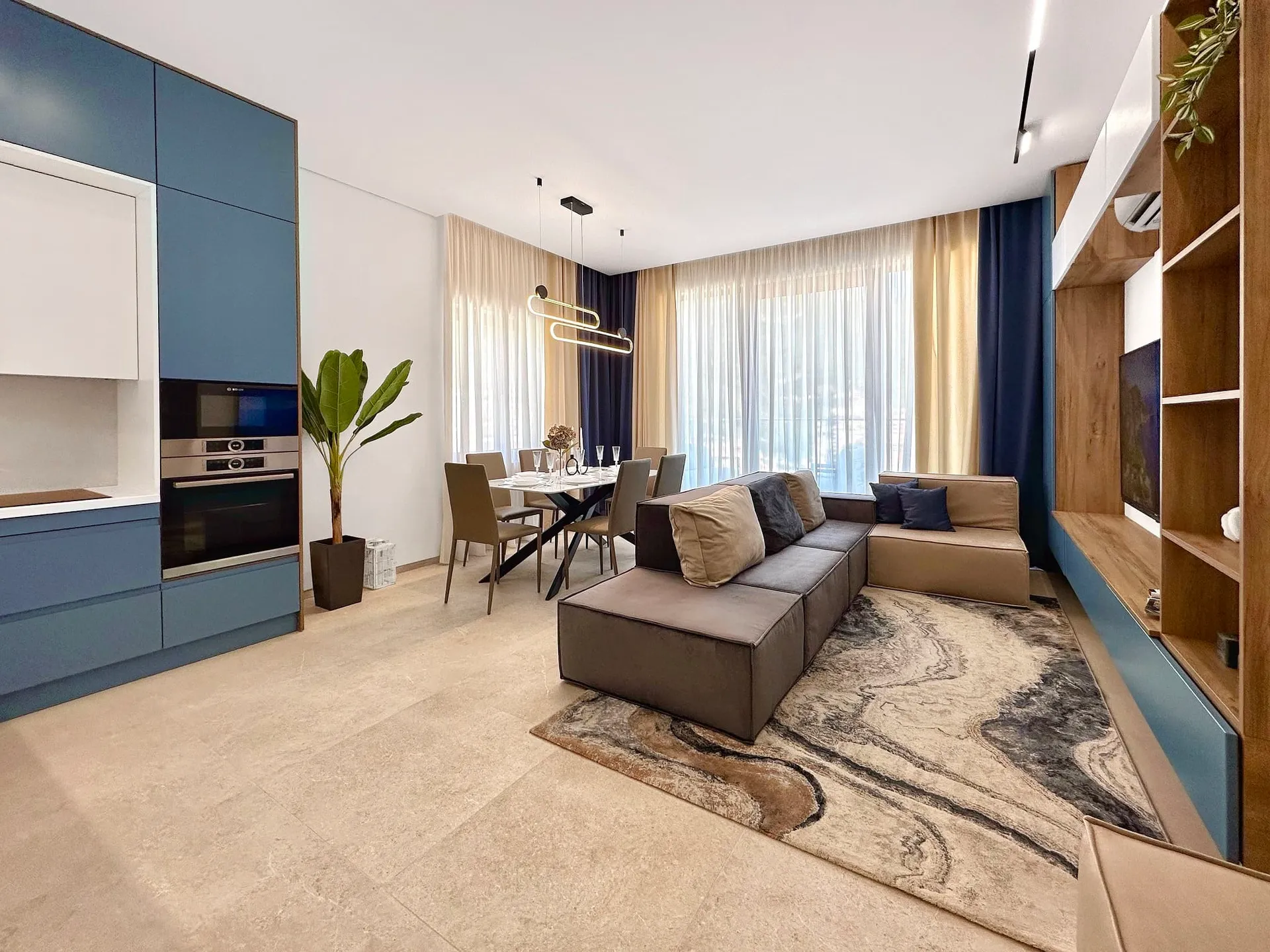 Modern open-plan living room with kitchen, grey sofa, dining table, and wooden TV wall unit.