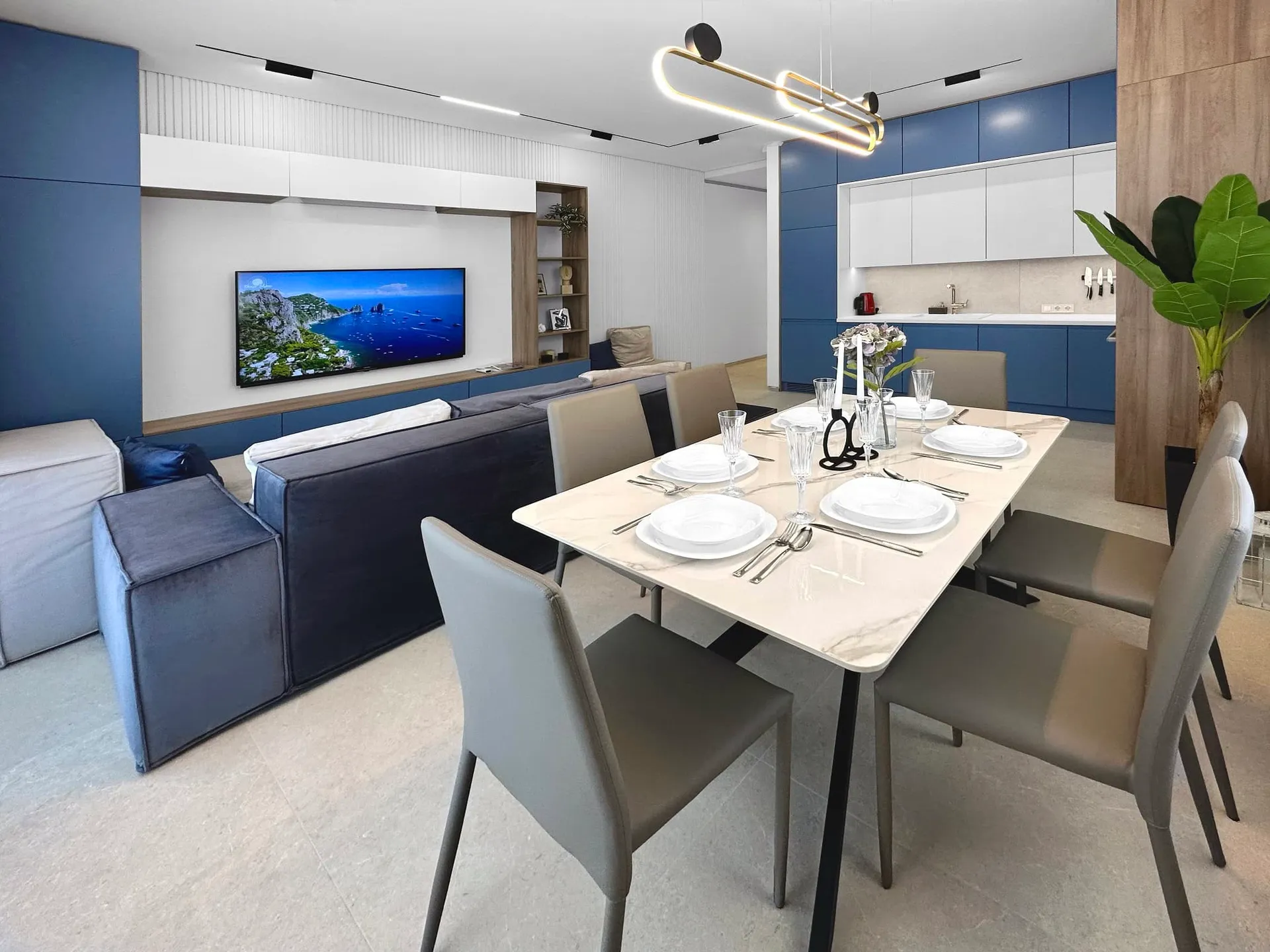 Living room featuring a blue sofa, wall-mounted TV, and a set dining table in the foreground with a kitchen visible behind.