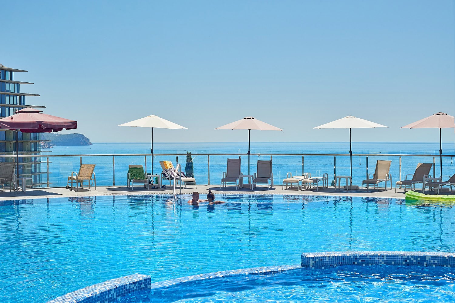 Outdoor infinity pool with a sea view at a residential complex in Becici, Montenegro, featuring sun loungers and umbrellas.