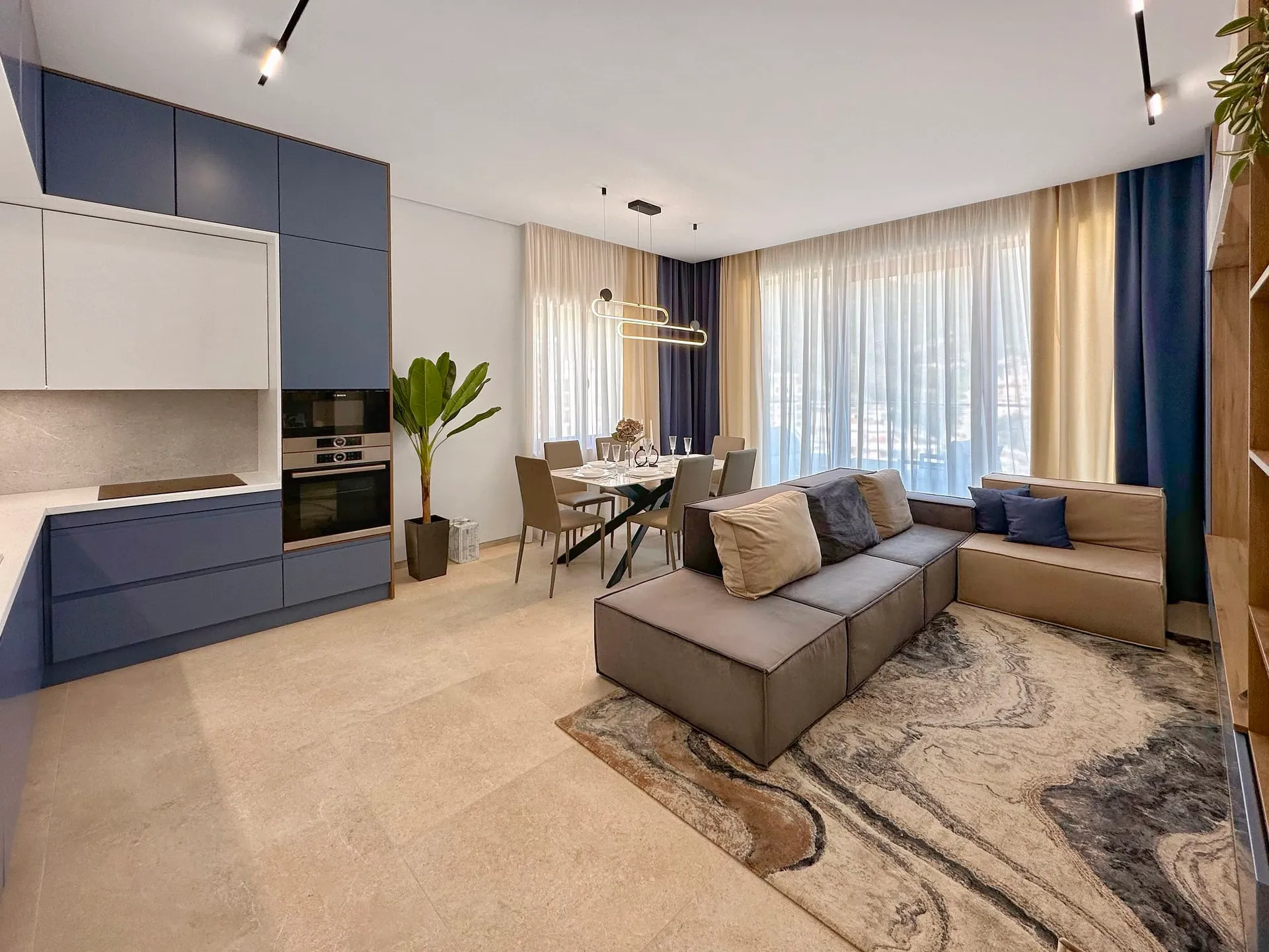 Modern open-plan living room with kitchen, grey sofa, dining table, and blue cabinetry.