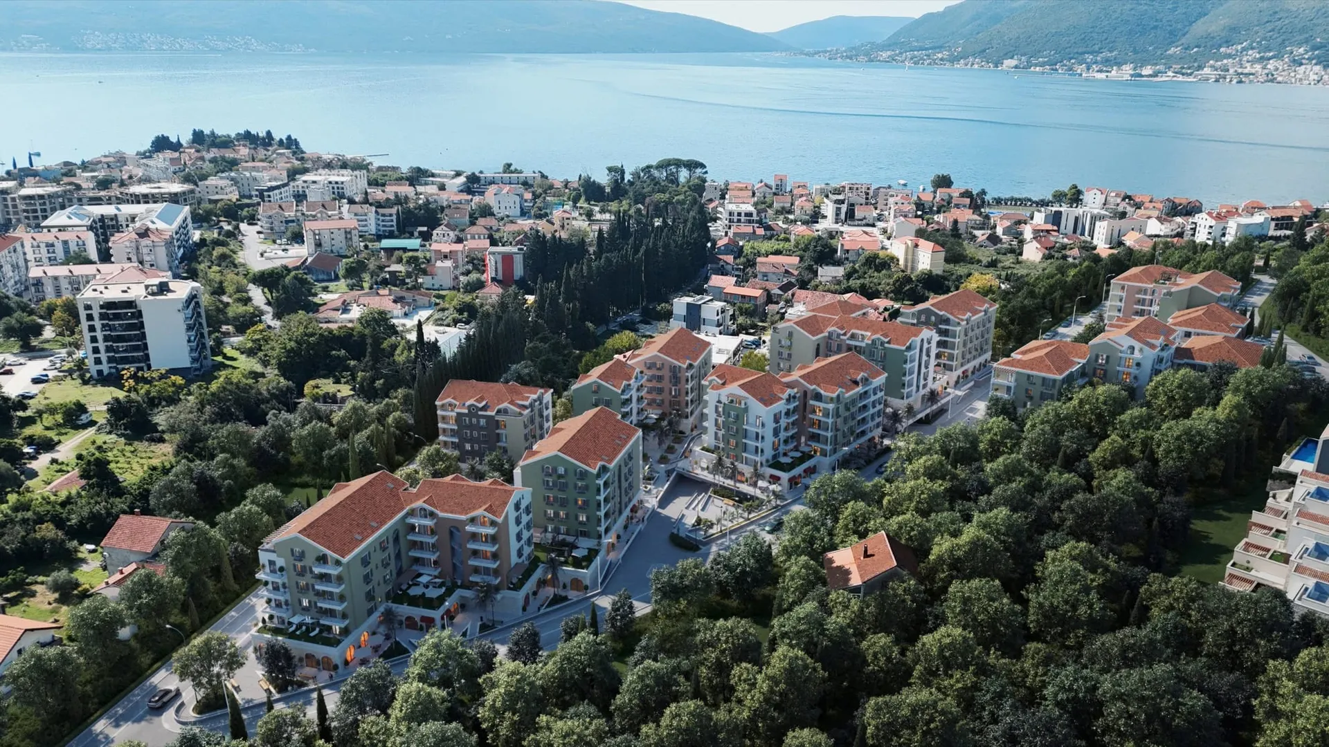 Aerial view of a residential complex with red roofs surrounded by trees near the sea and mountains.