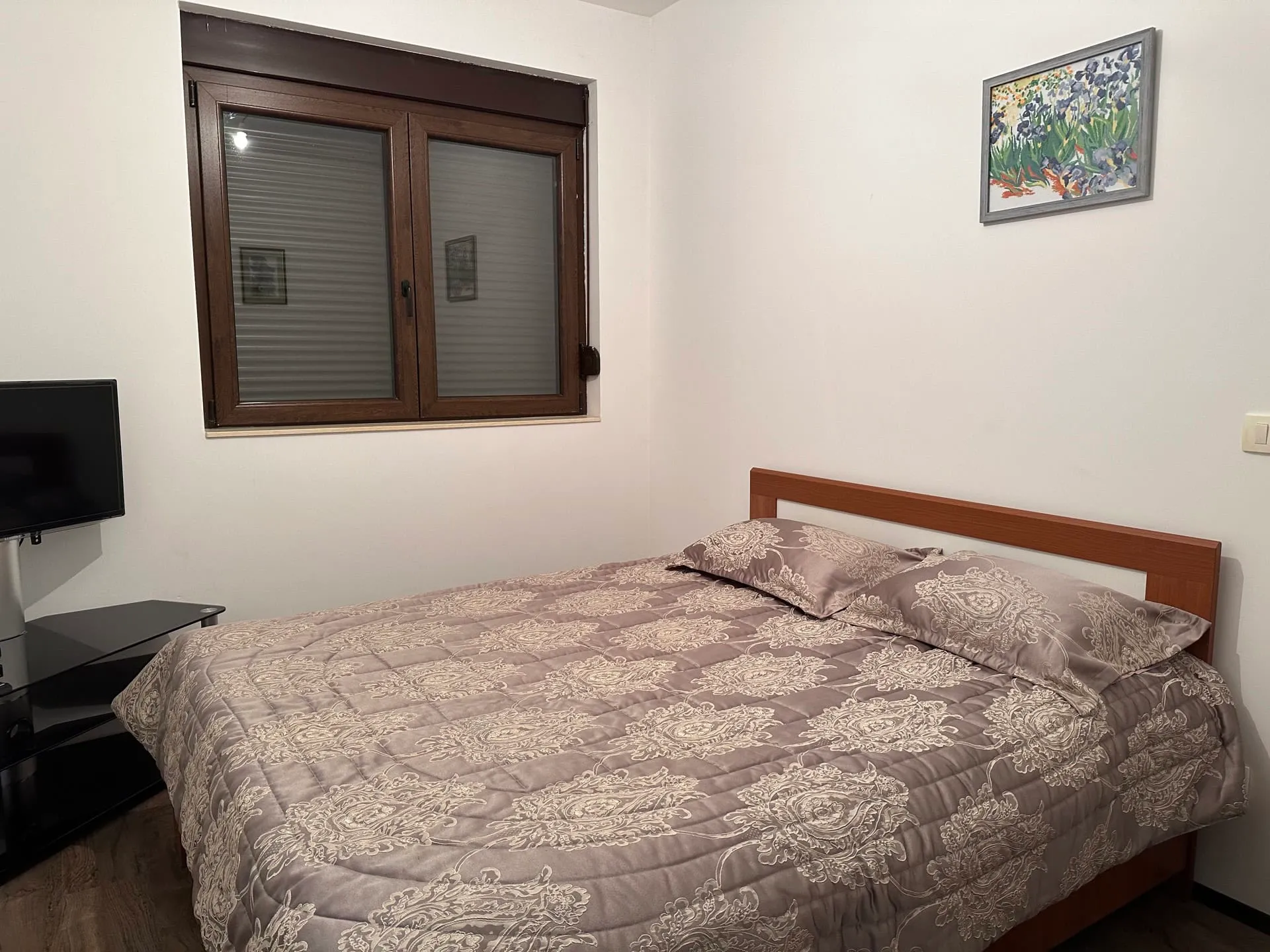 Bedroom featuring a wooden bed with grey patterned bedding, a TV on a stand, and a window with blinds.