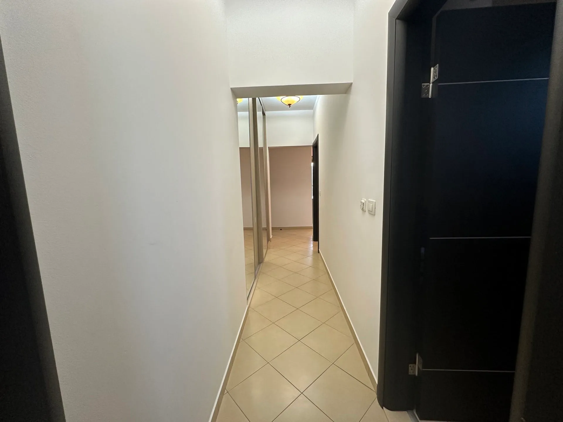 Narrow hallway with beige tiled floor, white walls, and a dark door on the right side.