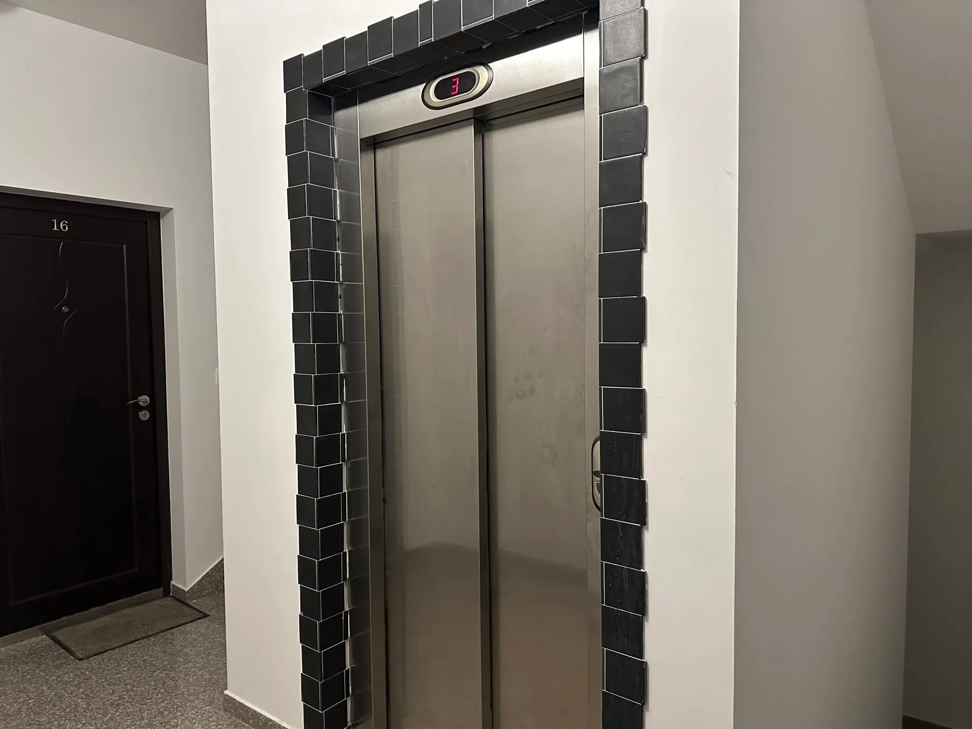 Elevator with metal doors framed in black tiles and a floor indicator next to a dark apartment door numbered 16.