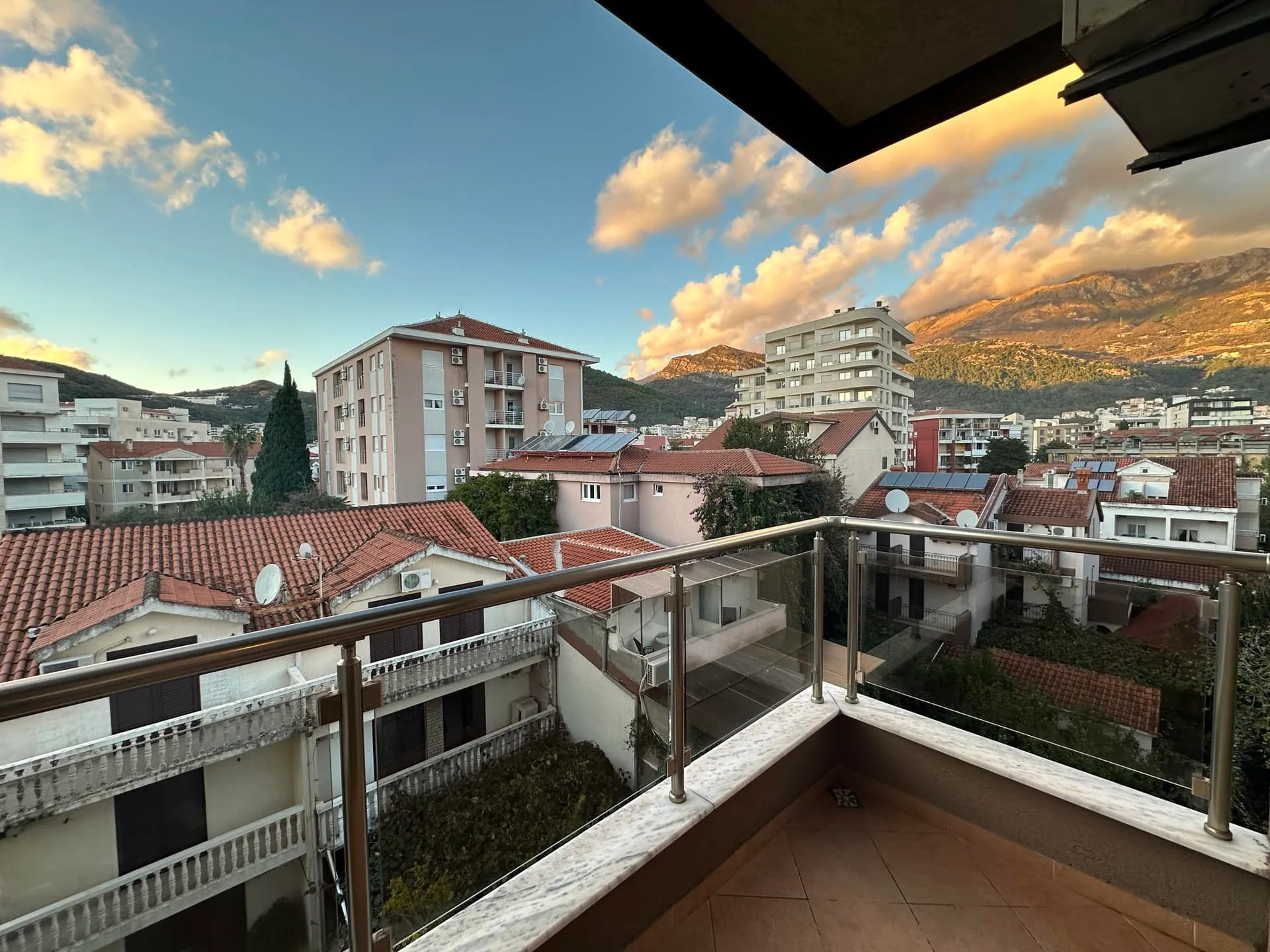 View from a balcony overlooking city rooftops and mountains. Features glass railing and tiled flooring.