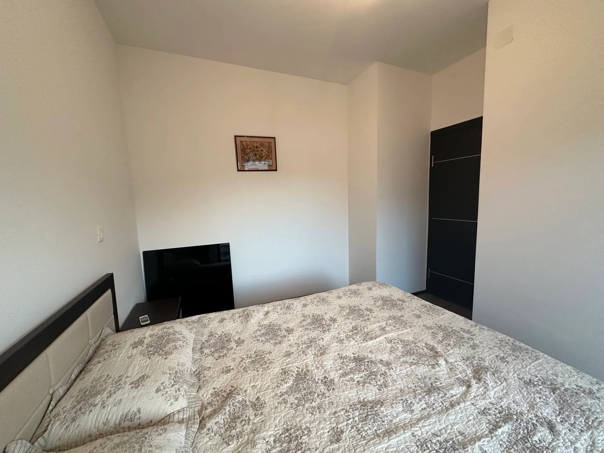 Bedroom featuring a double bed with floral cover, black TV, and dark door against white walls.
