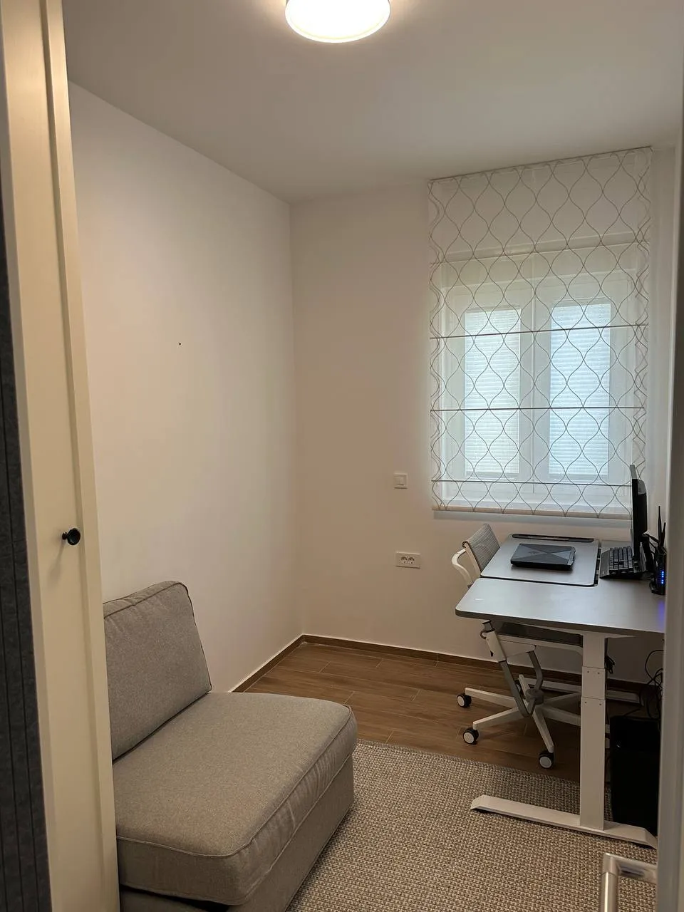 Small room featuring a standing desk with a computer and a grey sofa near the window.