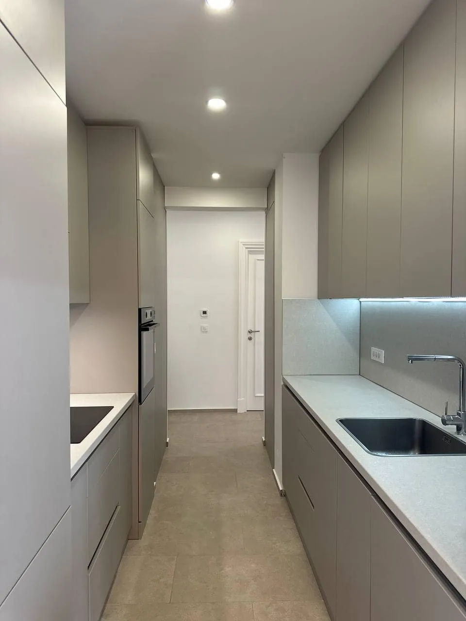 Narrow modern kitchen with grey cabinets, sink, cooktop, and built-in oven along a galley layout.