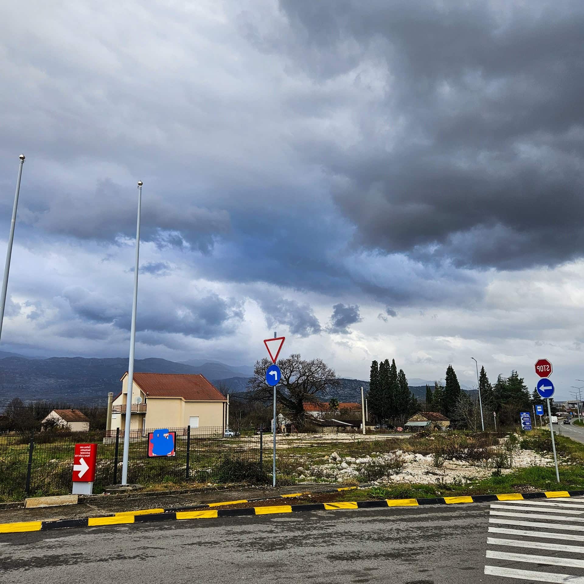 Street scene with road signs, empty land plot, and a house against mountains under stormy clouds.