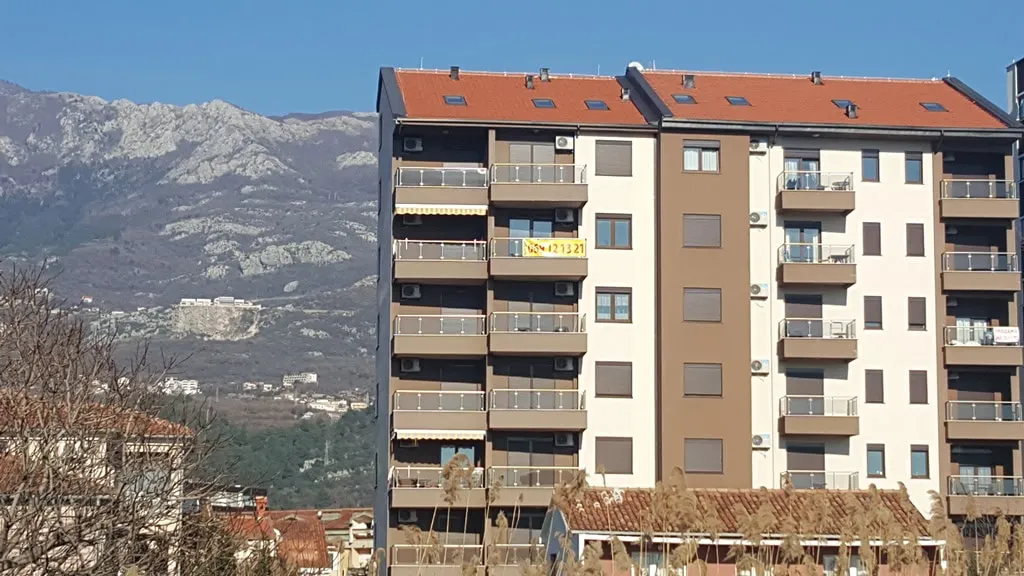 Exterior view of a multi-story residential building with balconies set against a backdrop of mountains and clear blue sky.