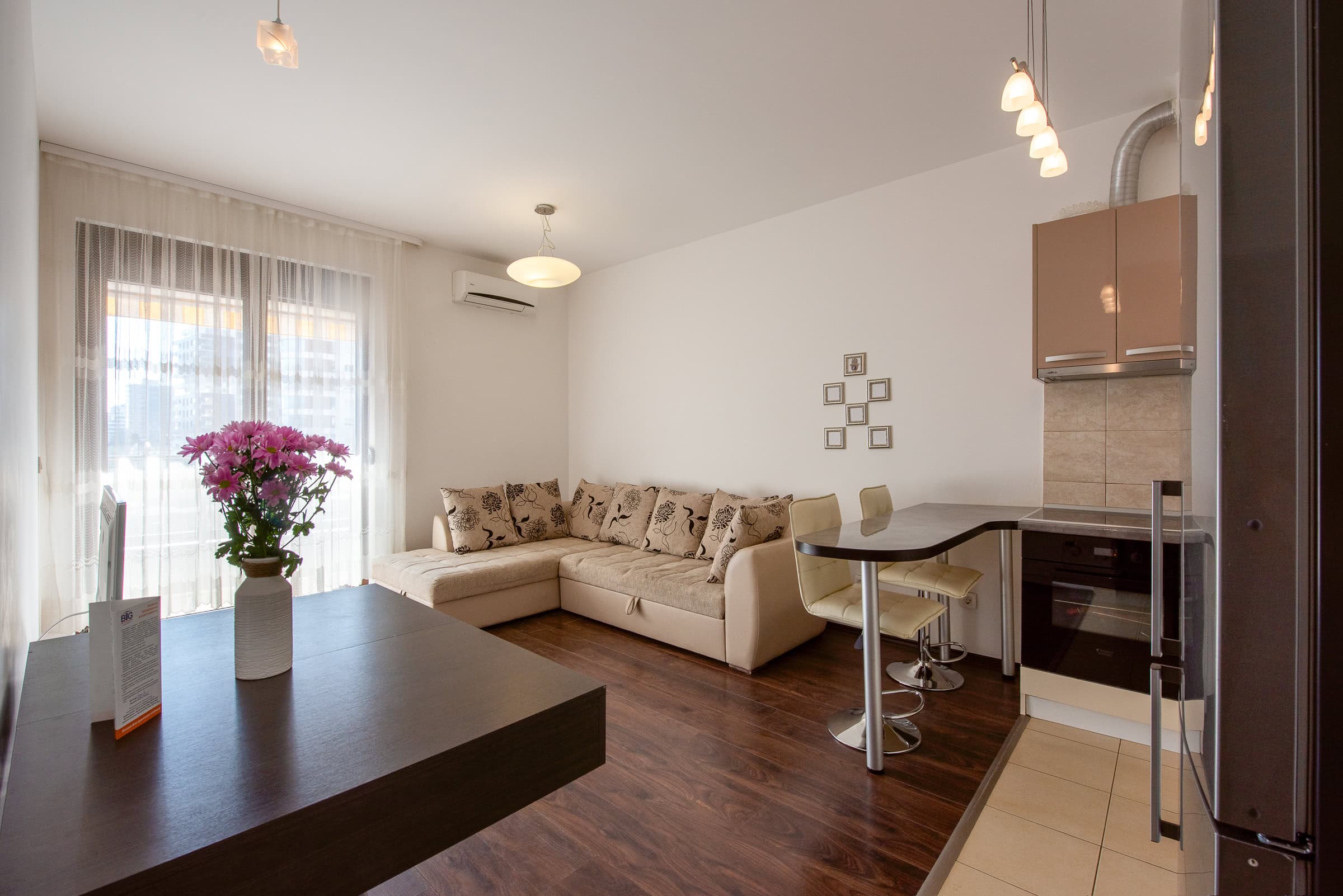 Living room with a beige sectional sofa, kitchen cabinets, bar stools, dark wood flooring, and a window with sheer curtains.