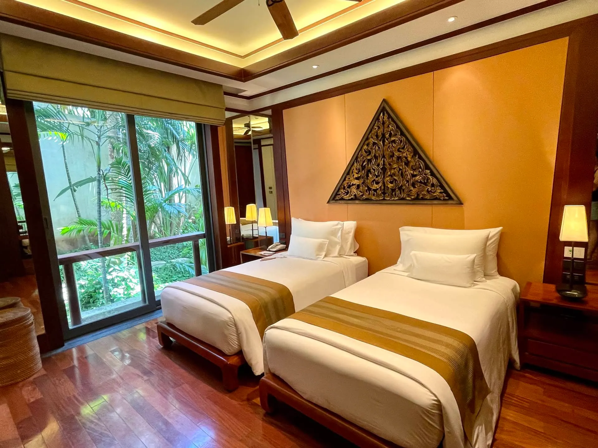 Two single beds with white linens and wooden frames facing a large window overlooking tropical greenery.