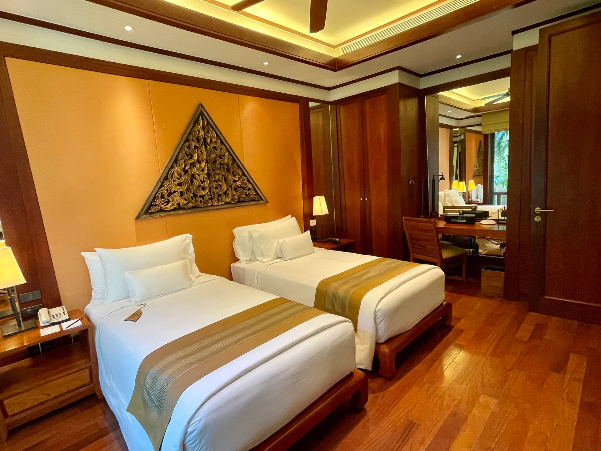 Two single beds with white linens, wooden flooring, decorative wall panel, and a desk area visible in the room.