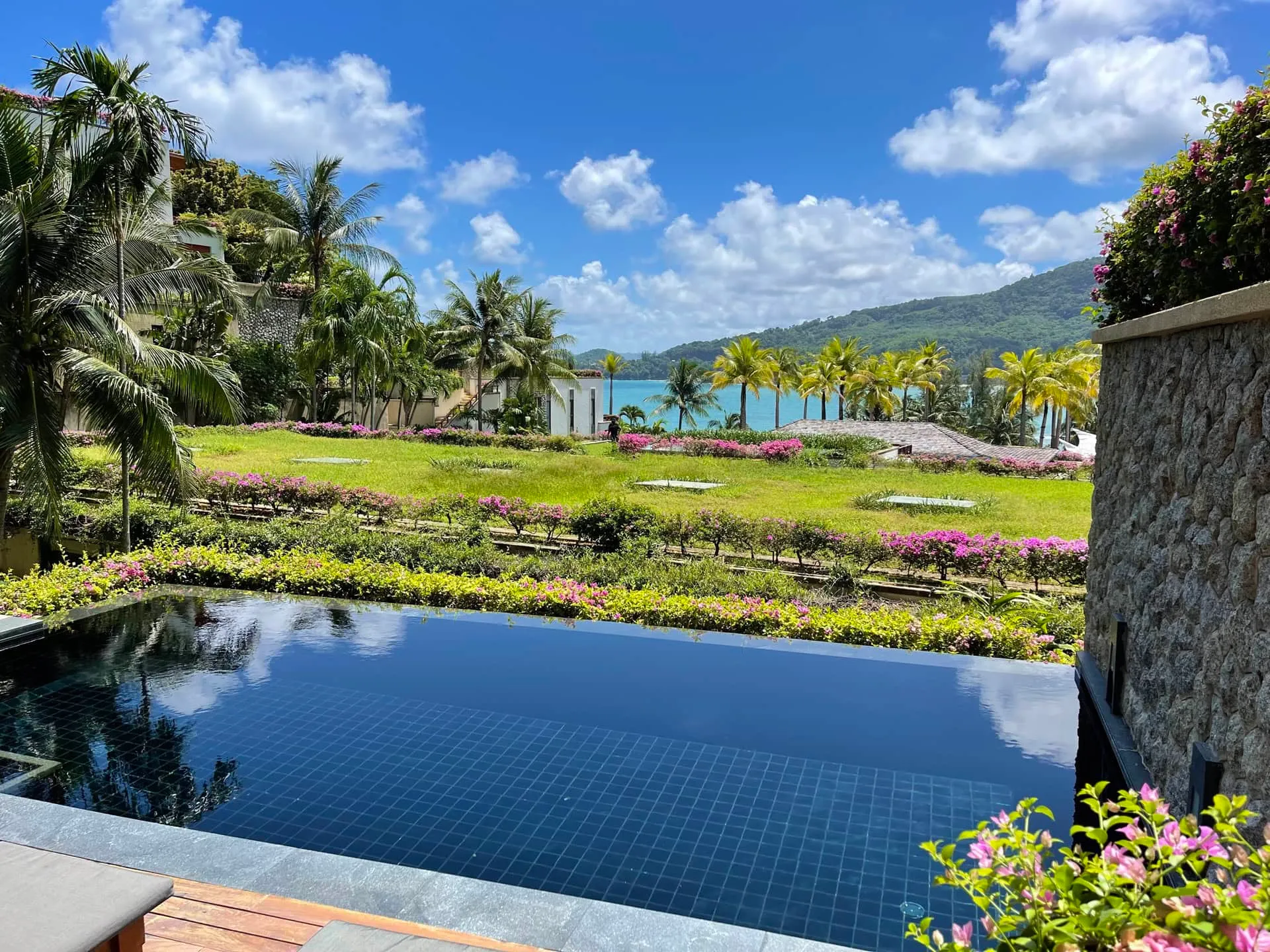 Dark tiled pool overlooking a tropical garden with palm trees, flowers, and the ocean.