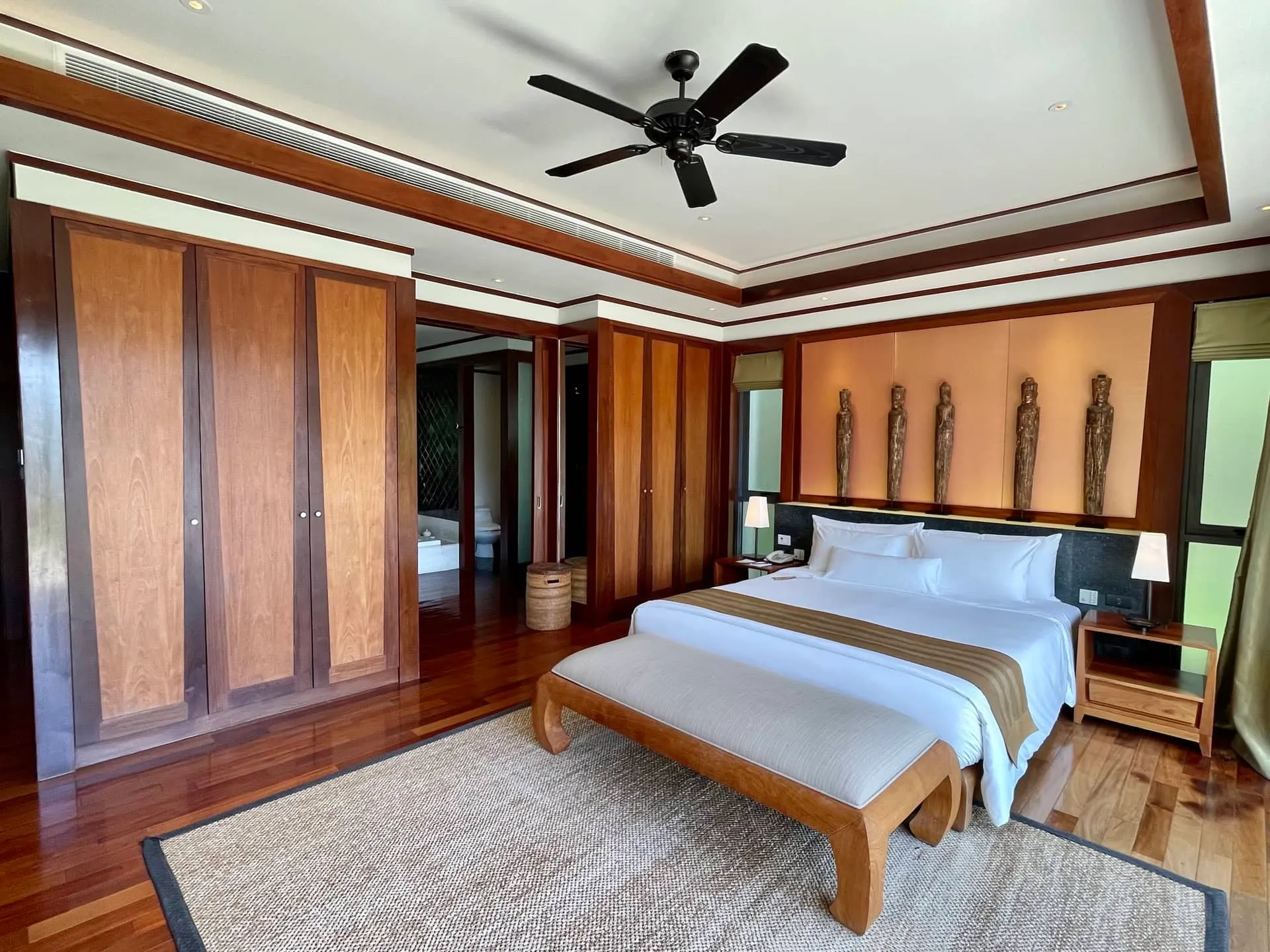 Bedroom featuring a large wooden bed, built-in wardrobes, ceiling fan, and decorative statues above the headboard on wood flooring.