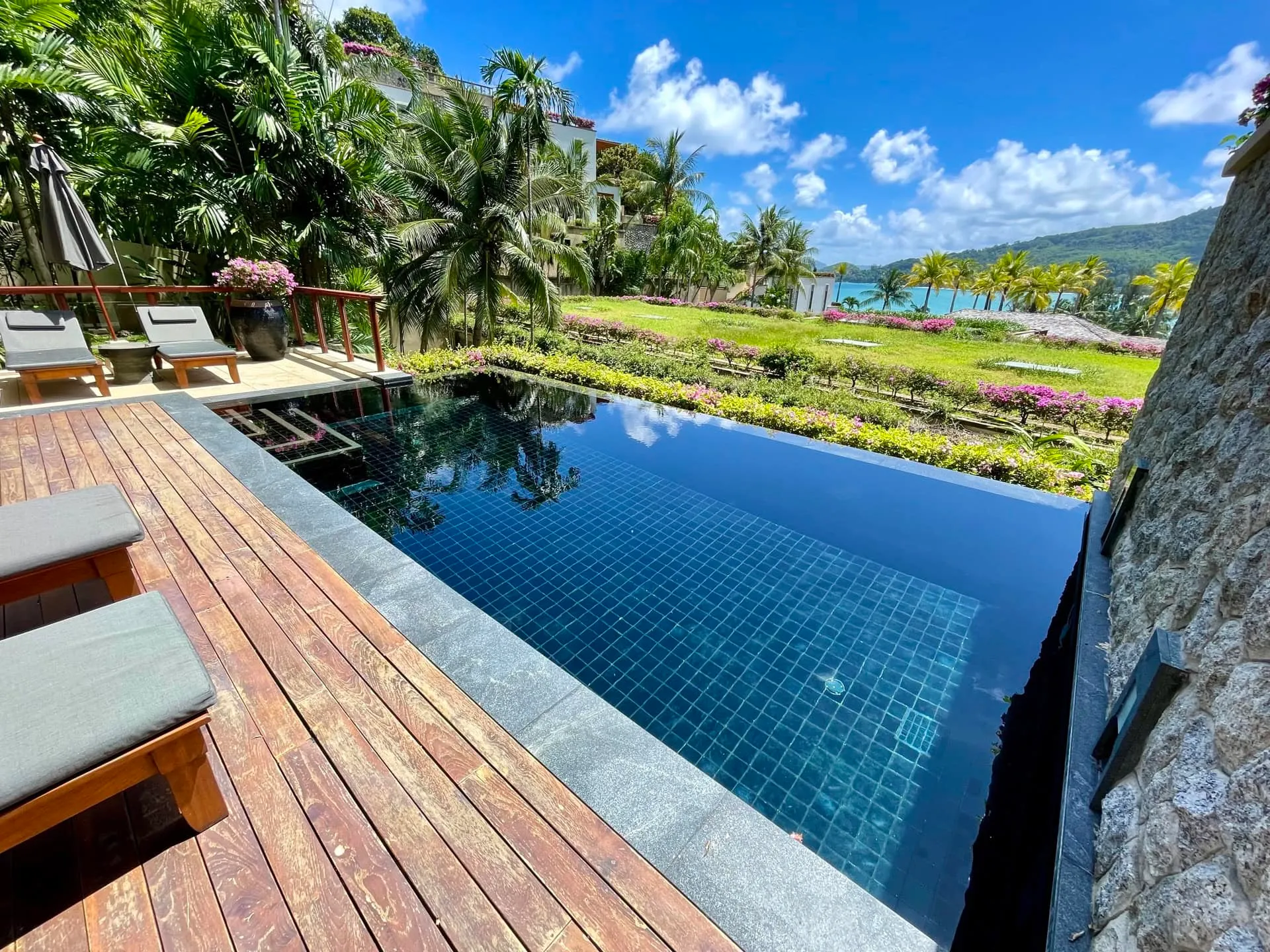 Infinity pool with wooden deck and loungers overlooking tropical garden and ocean view.