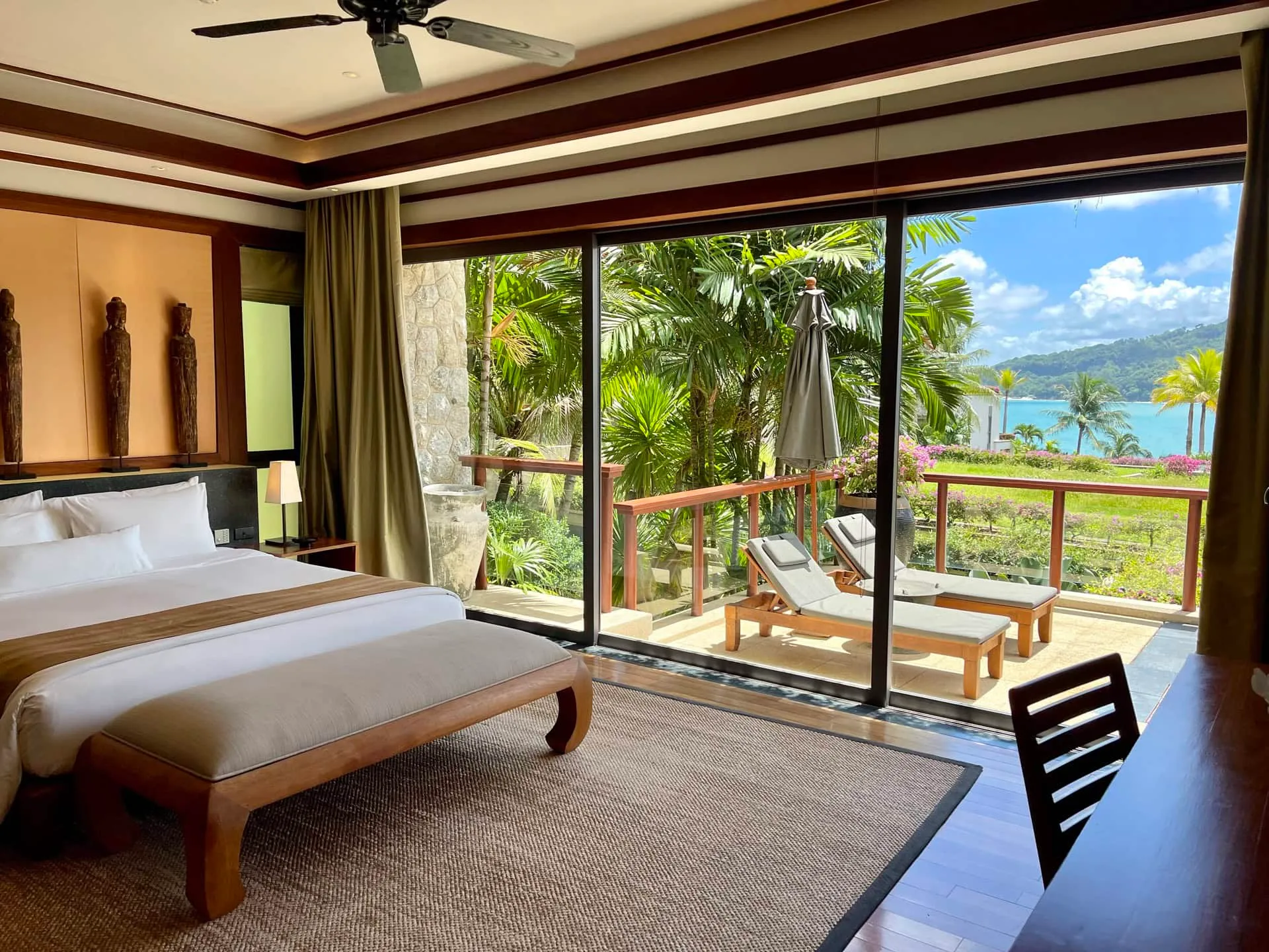 Bedroom featuring a large bed, wood ceiling, and sliding glass doors opening to a balcony with ocean and garden views.