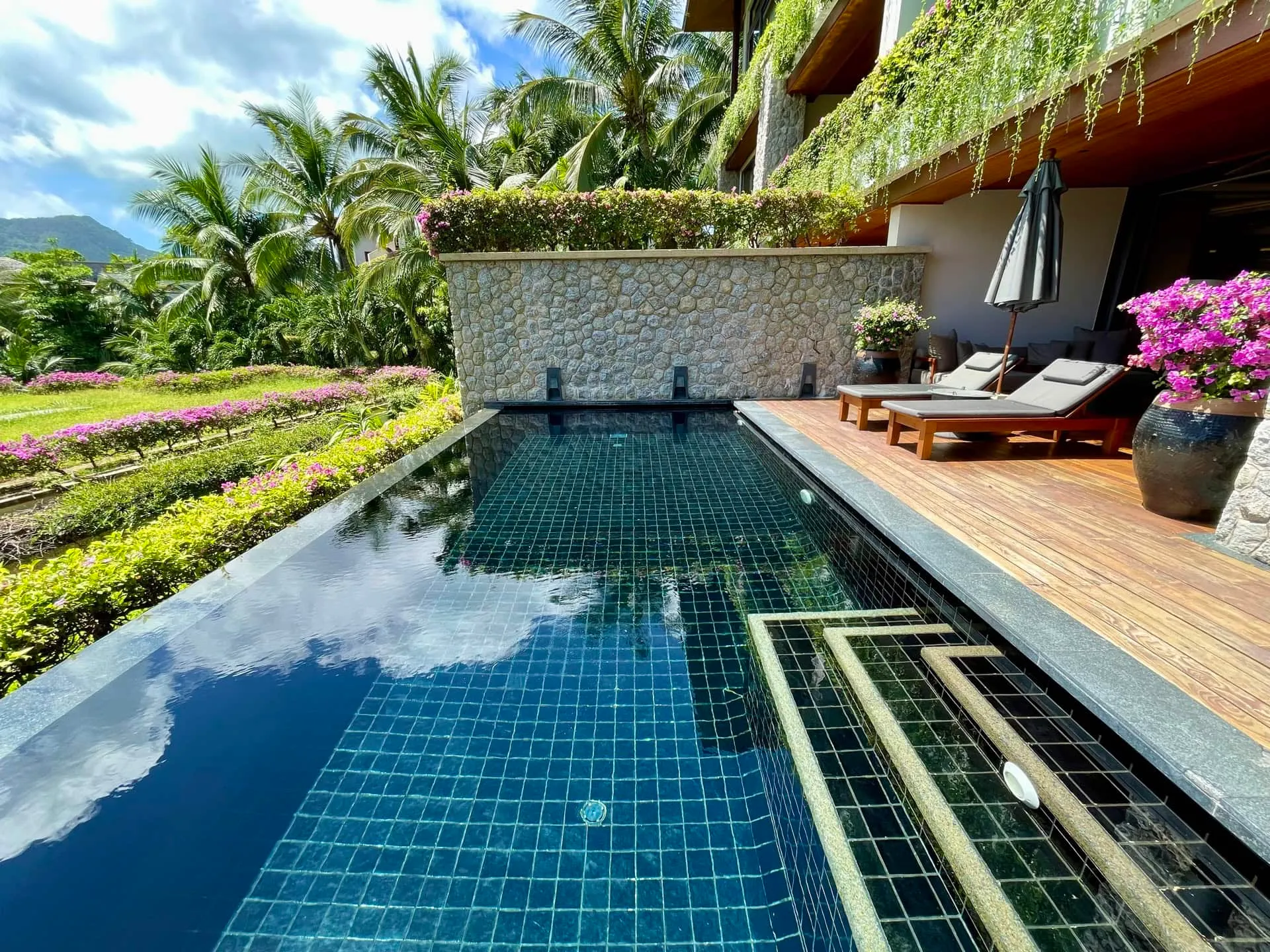 Long blue tiled swimming pool with wooden deck, lounge chairs, and tropical garden scenery.