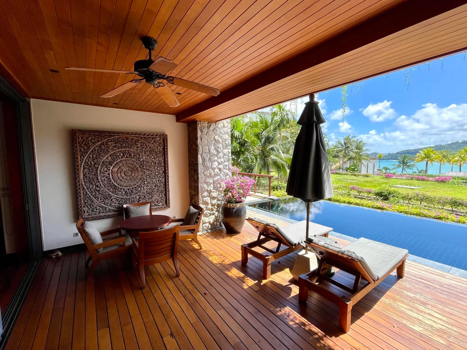 Wooden deck with dining set, sunbeds, and umbrella next to an infinity pool overlooking the ocean.