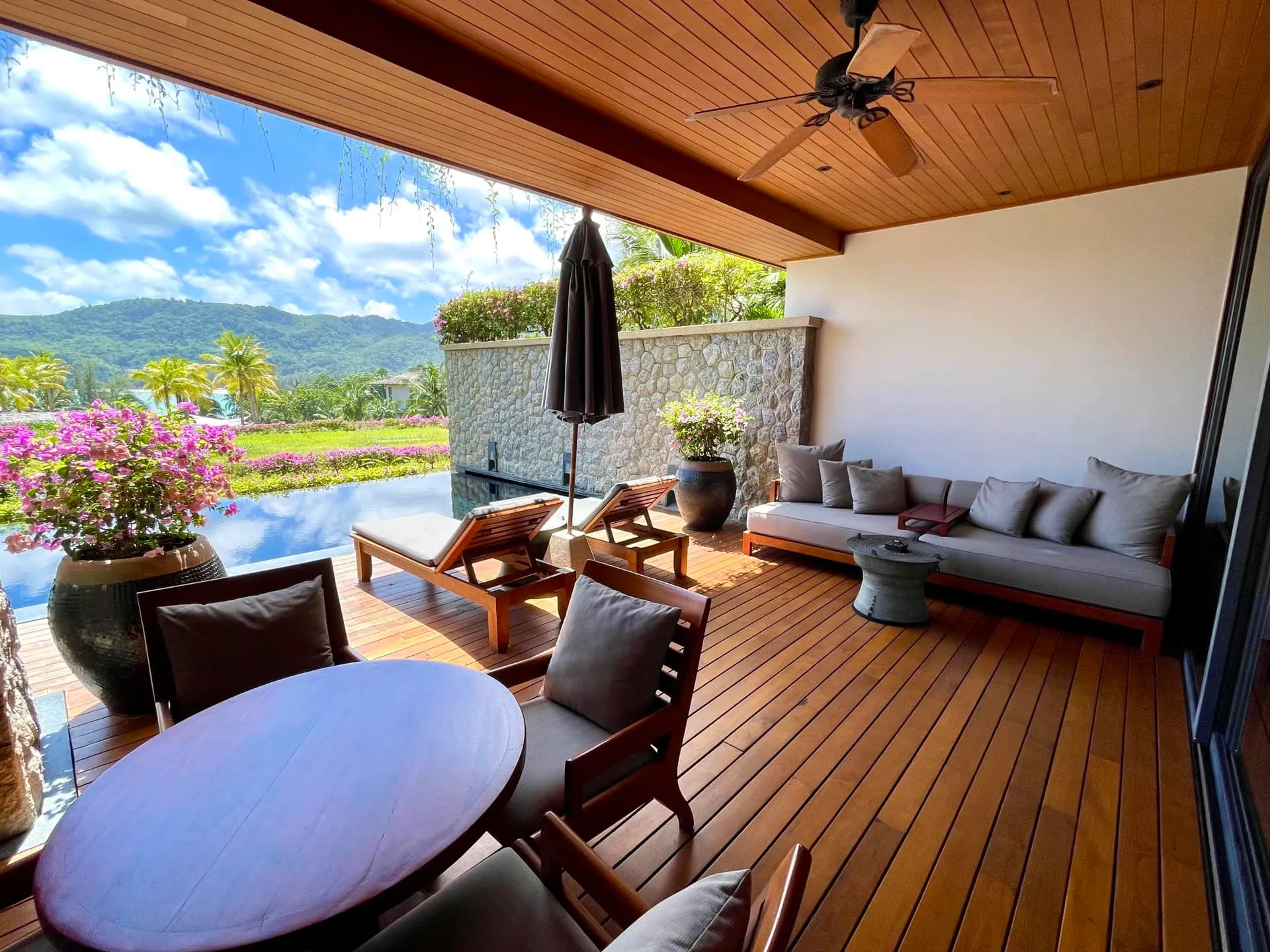 Wooden terrace with sofa, table, and sunbeds by a pool overlooking mountains and ocean.