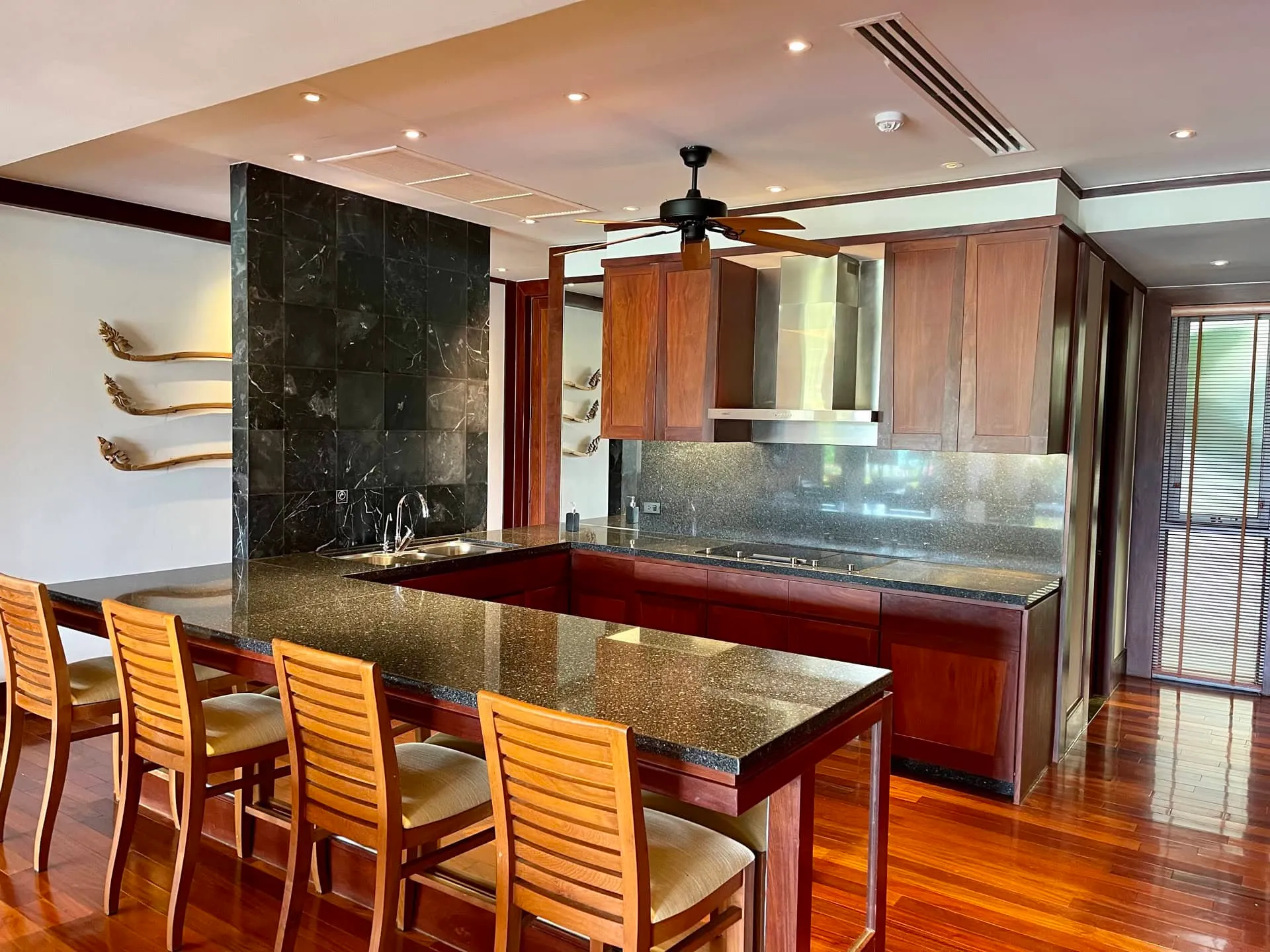 Modern kitchen featuring wooden cabinetry, dark granite countertops, a dining island with chairs, and a stainless steel hood.