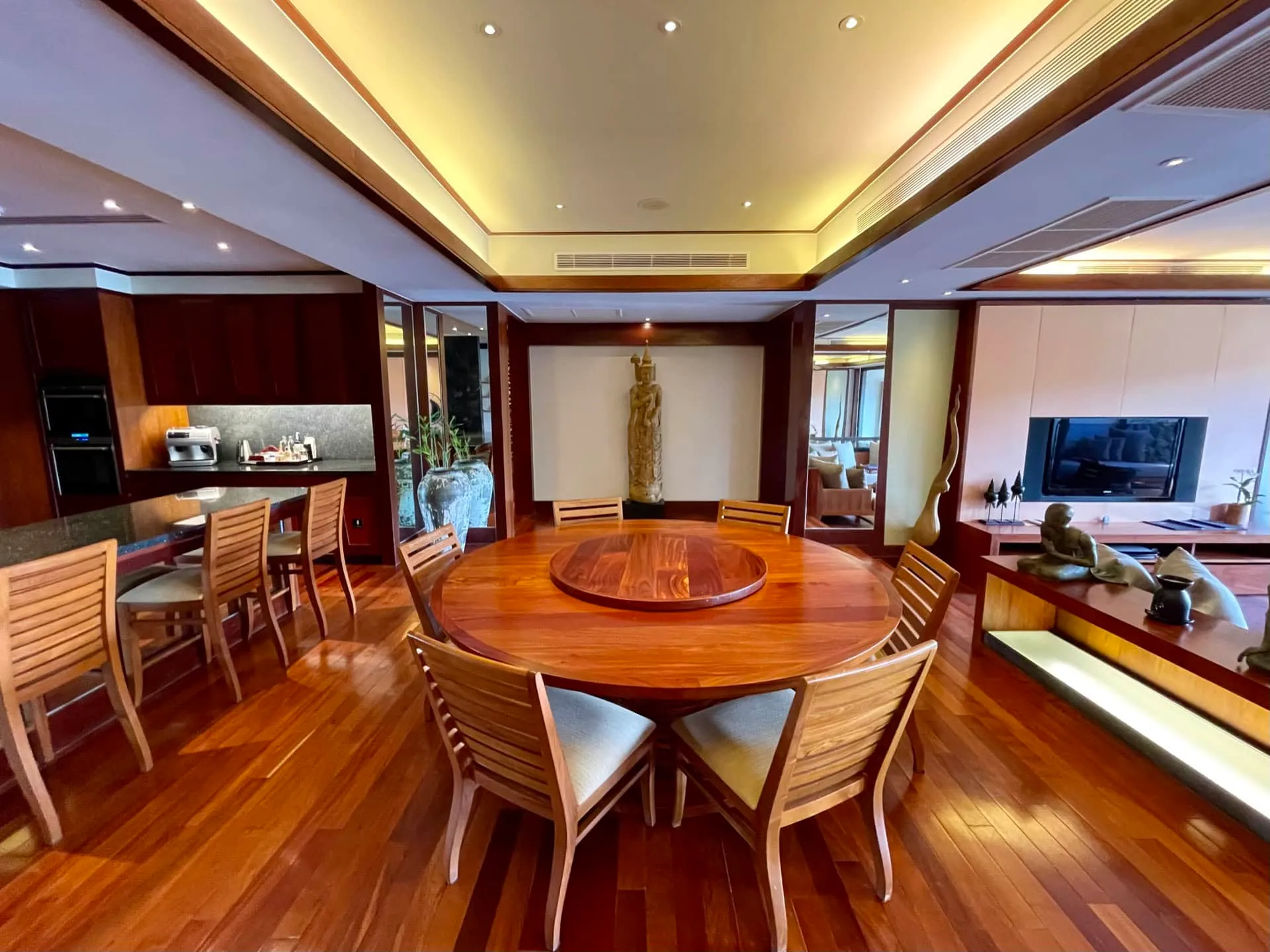 Dining area with a round wooden table, adjacent kitchen counter visible, and living room with TV in background.