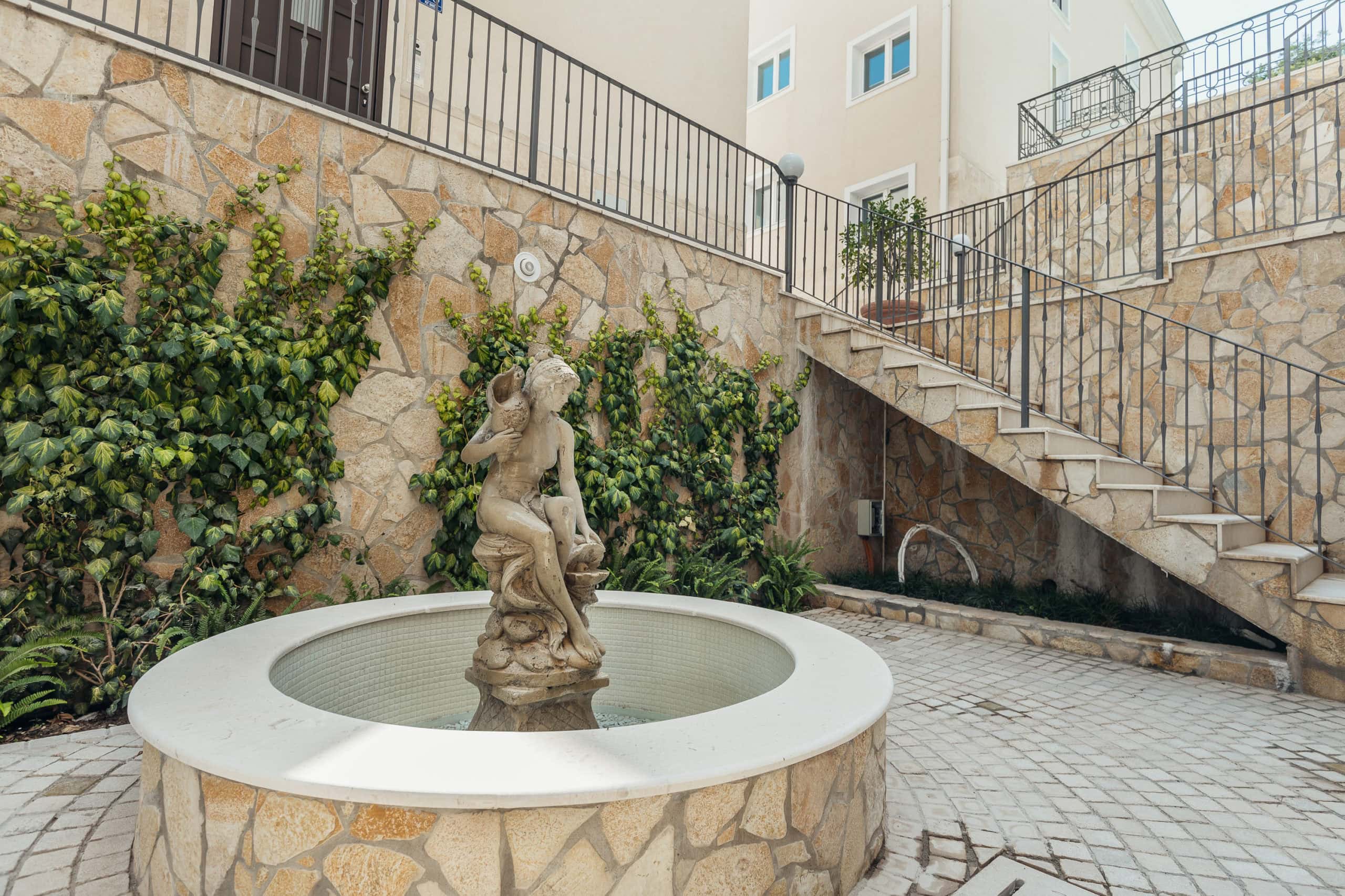 Courtyard of a residential complex in Kumbor, Montenegro: stone wall finishes, a fountain with a statue, and stairs leading to the apartments.
