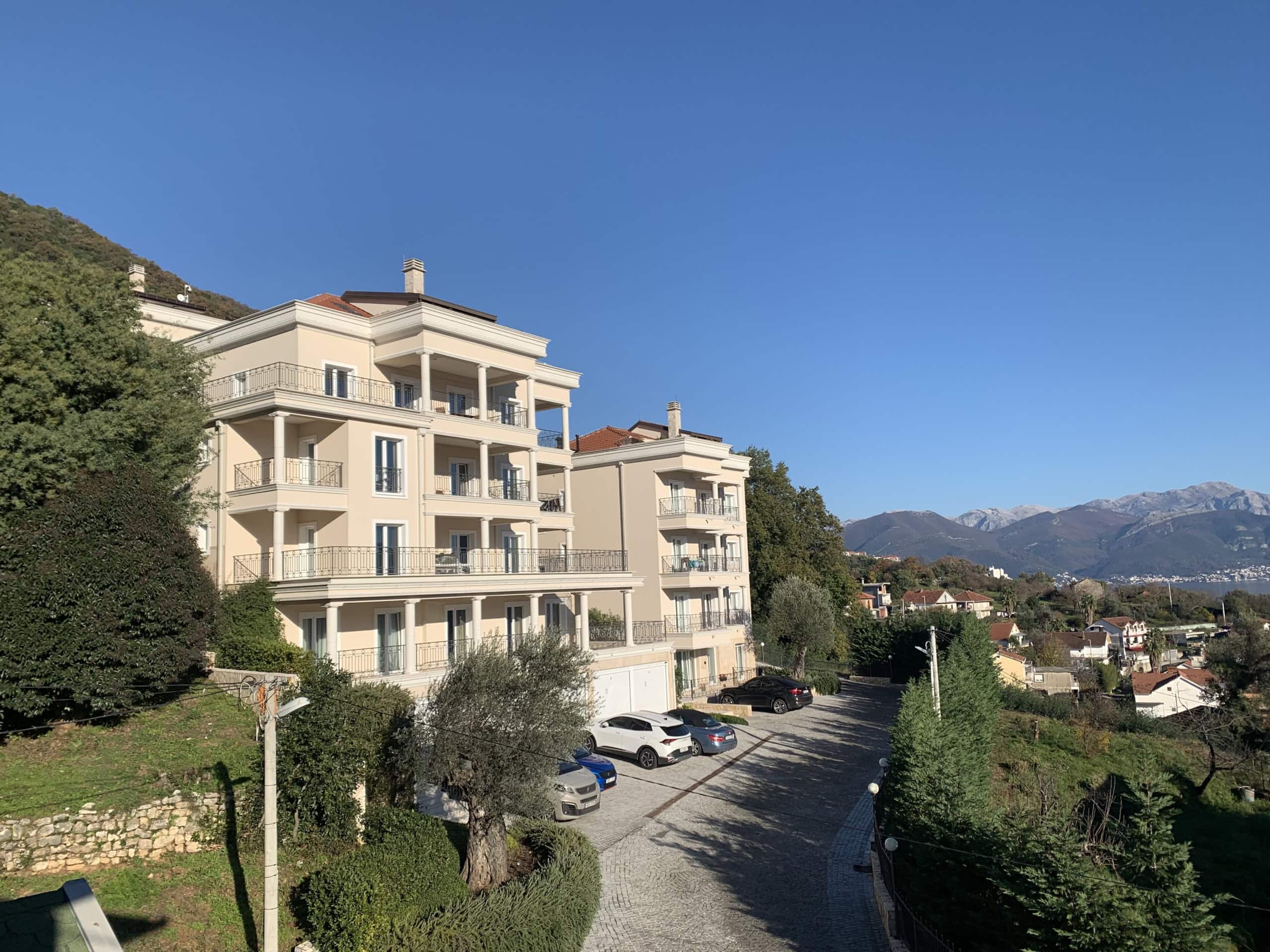 Modern residential complex with terraces in Kumbor, Montenegro: view of the building, parking area, and mountains on a sunny day.
