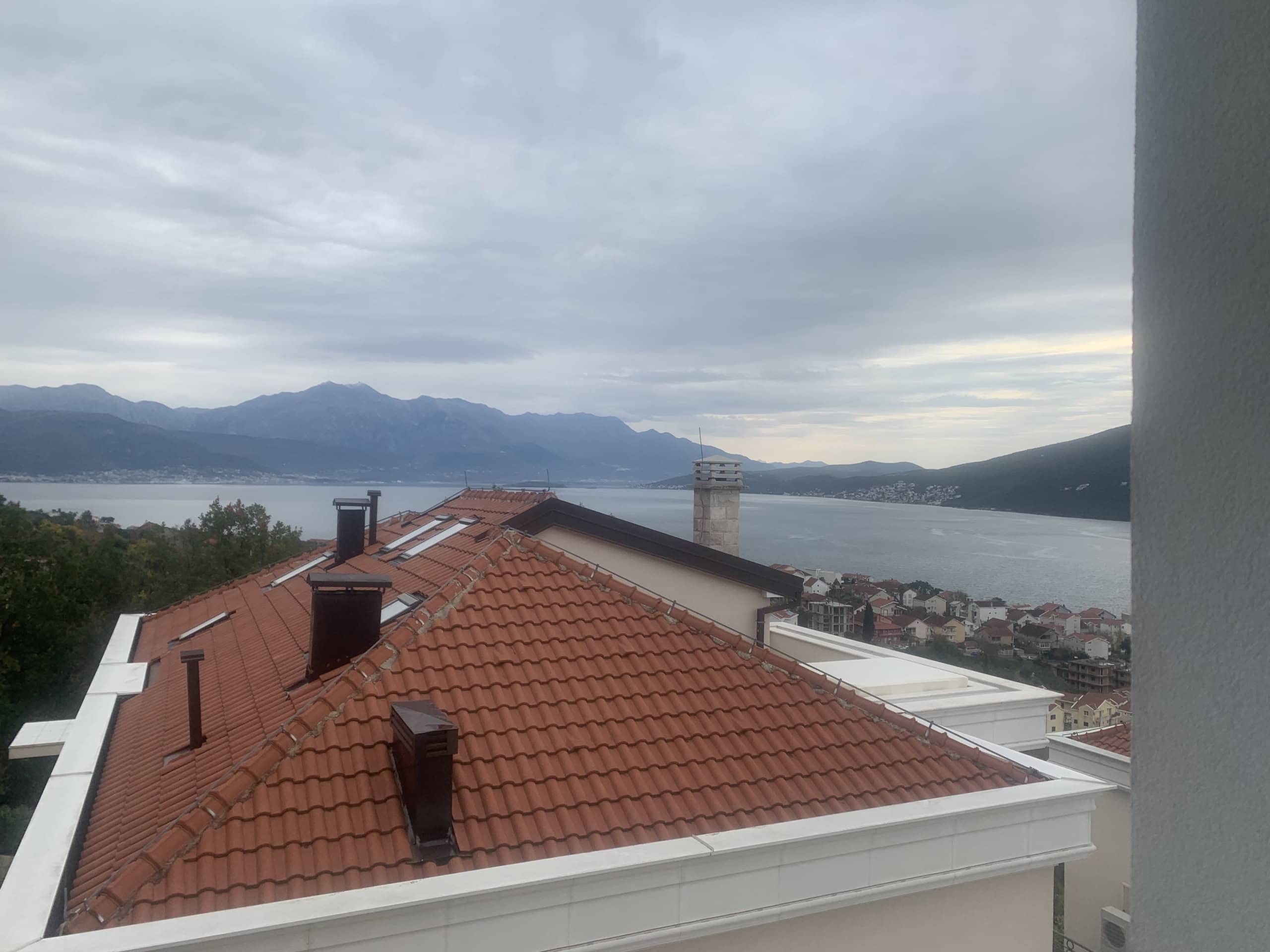 View from the balcony of a residential complex in Kumbor, Montenegro: a panorama of the sea, mountains, and the red-tiled roofs of houses.