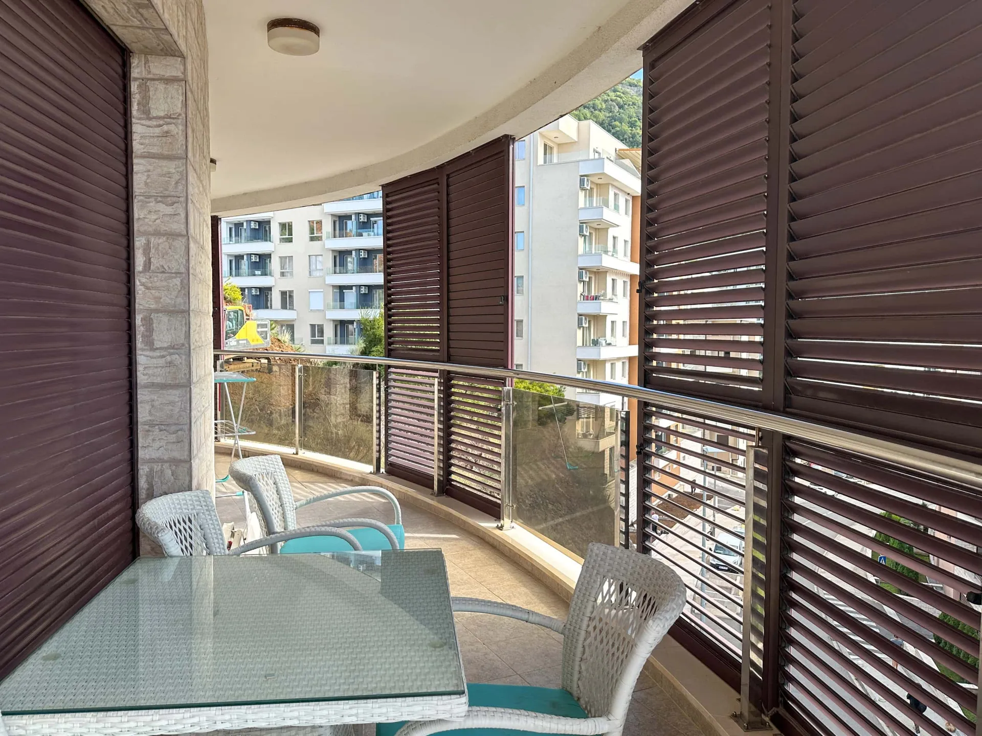 Balcony with white wicker chairs, glass table, brown louvered shutters, and a view of nearby apartment buildings.