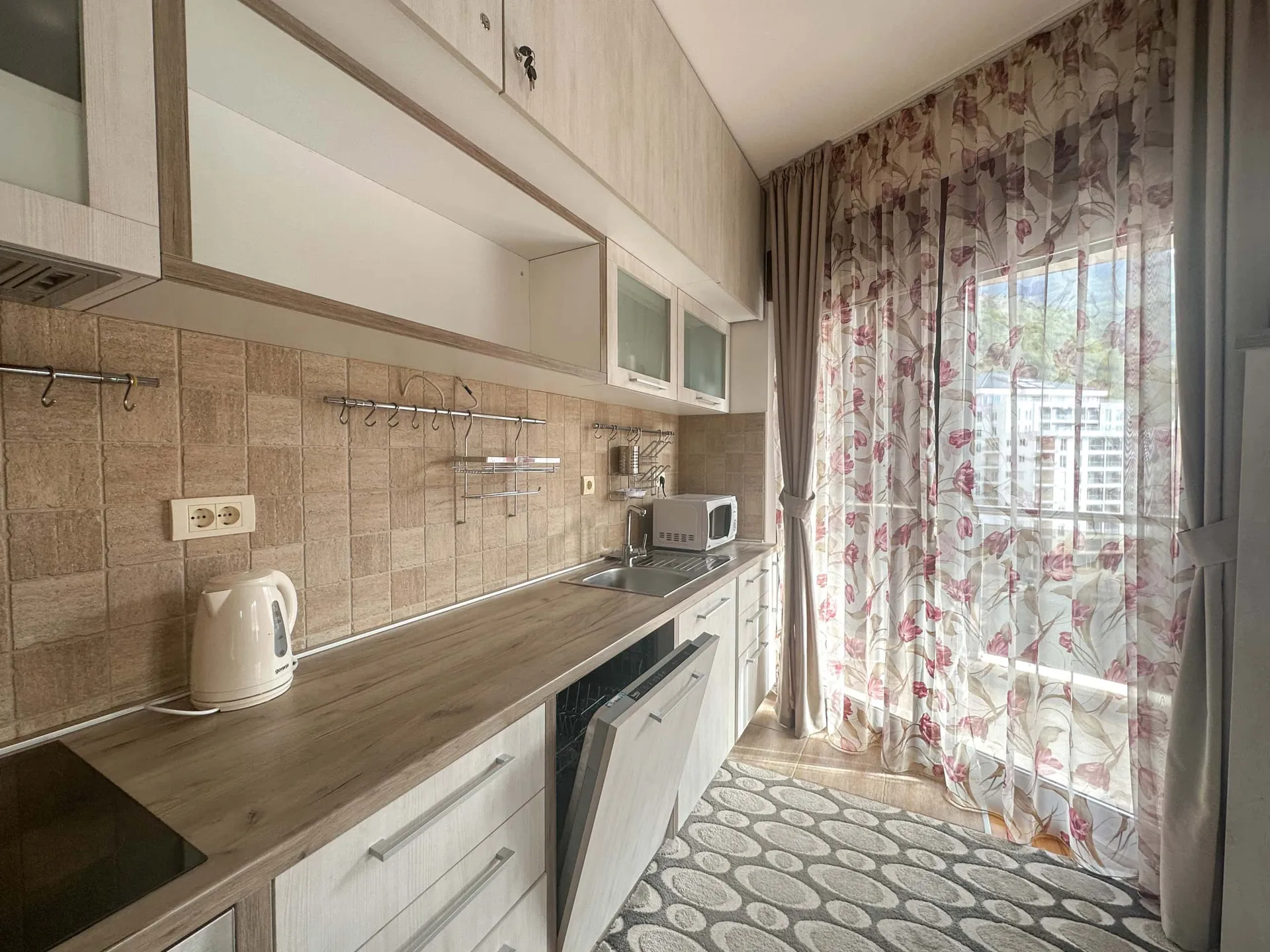Kitchen with light cabinets, wood countertop, kettle, microwave, sink near a window with floral curtains and a patterned rug.