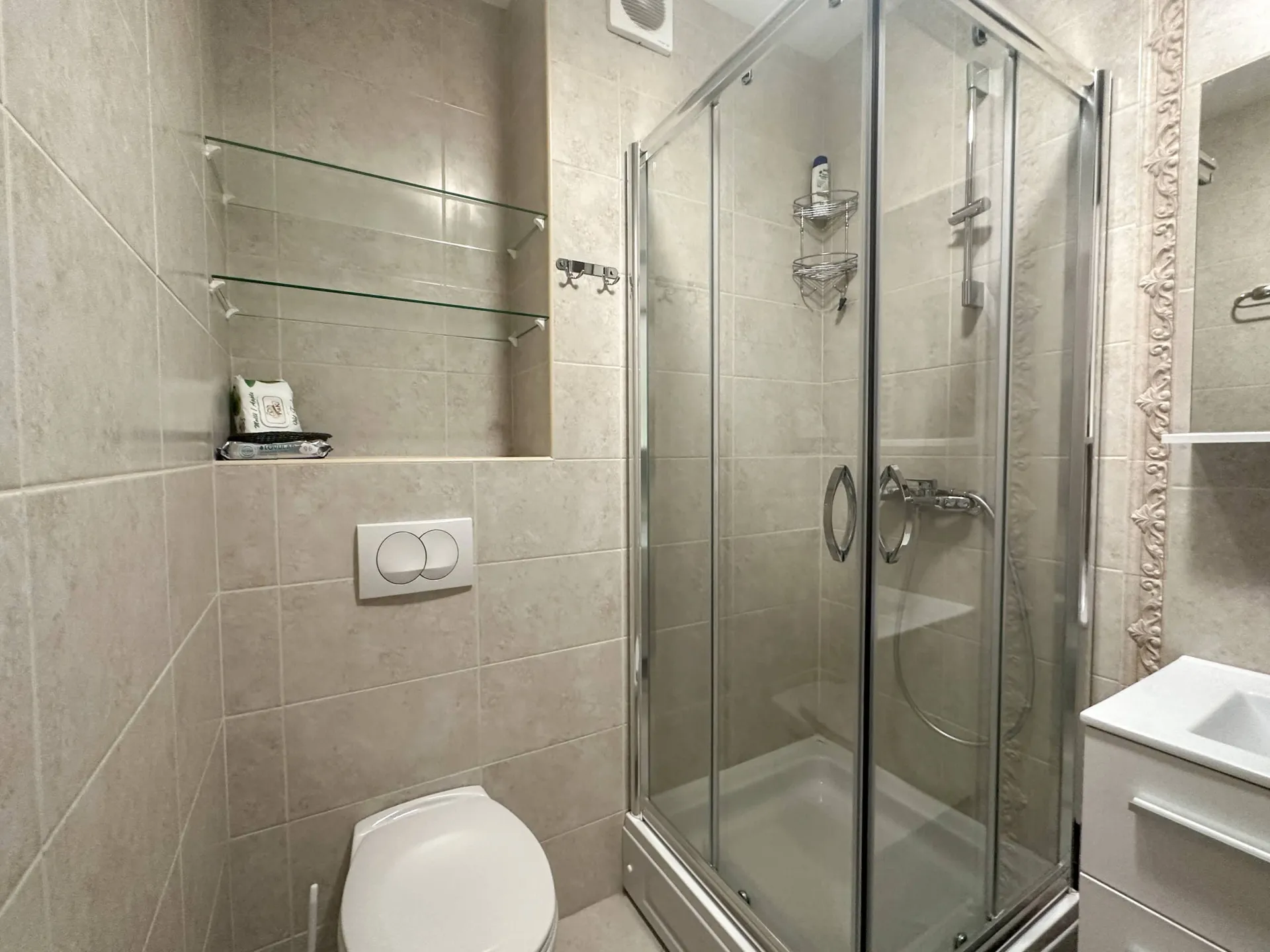 Bathroom featuring a glass shower enclosure, toilet, wall shelves, and beige tiled walls.