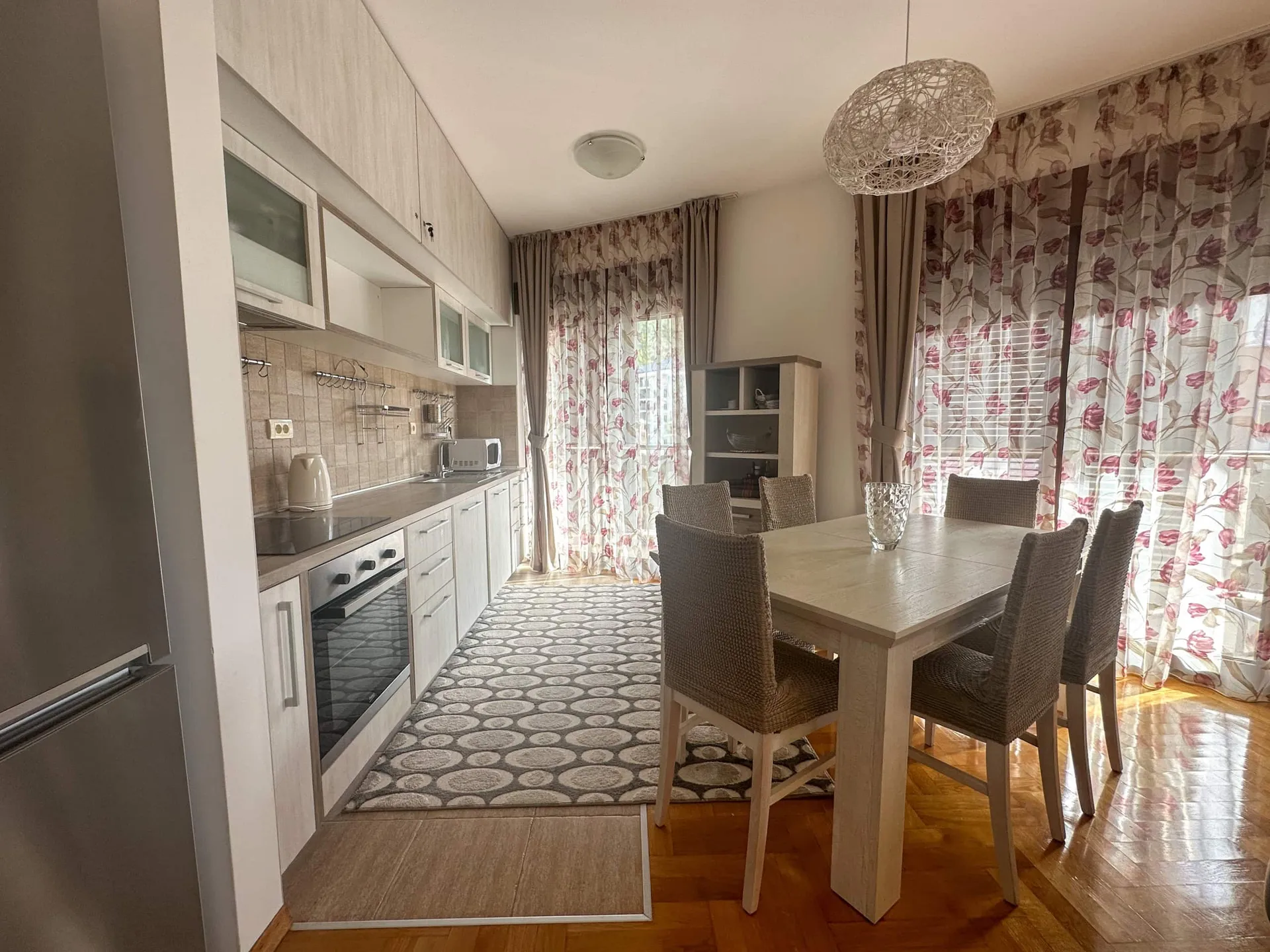 Kitchen with light cabinets, oven, fridge, and dining table near windows covered by floral curtains.
