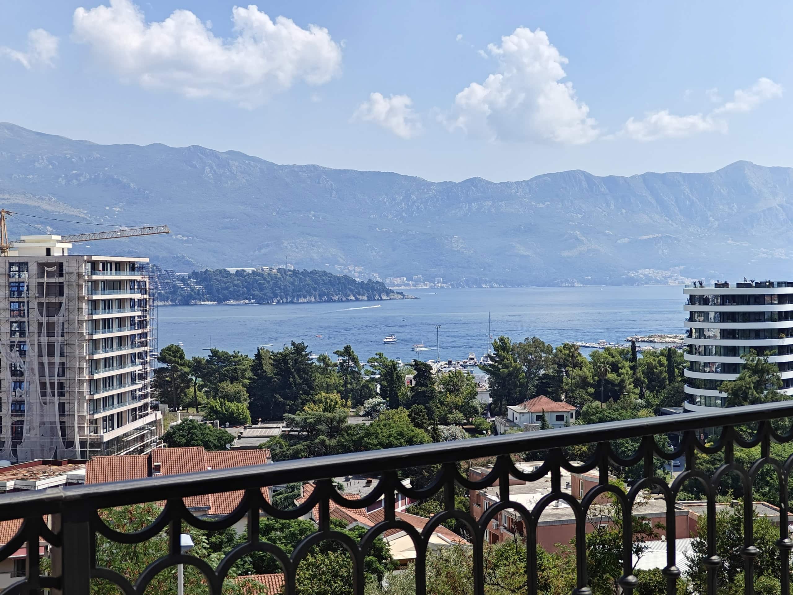 View from the apartment balcony in Budva, Montenegro: a panorama of the Adriatic Sea, mountains, yachts, and the city’s modern development.