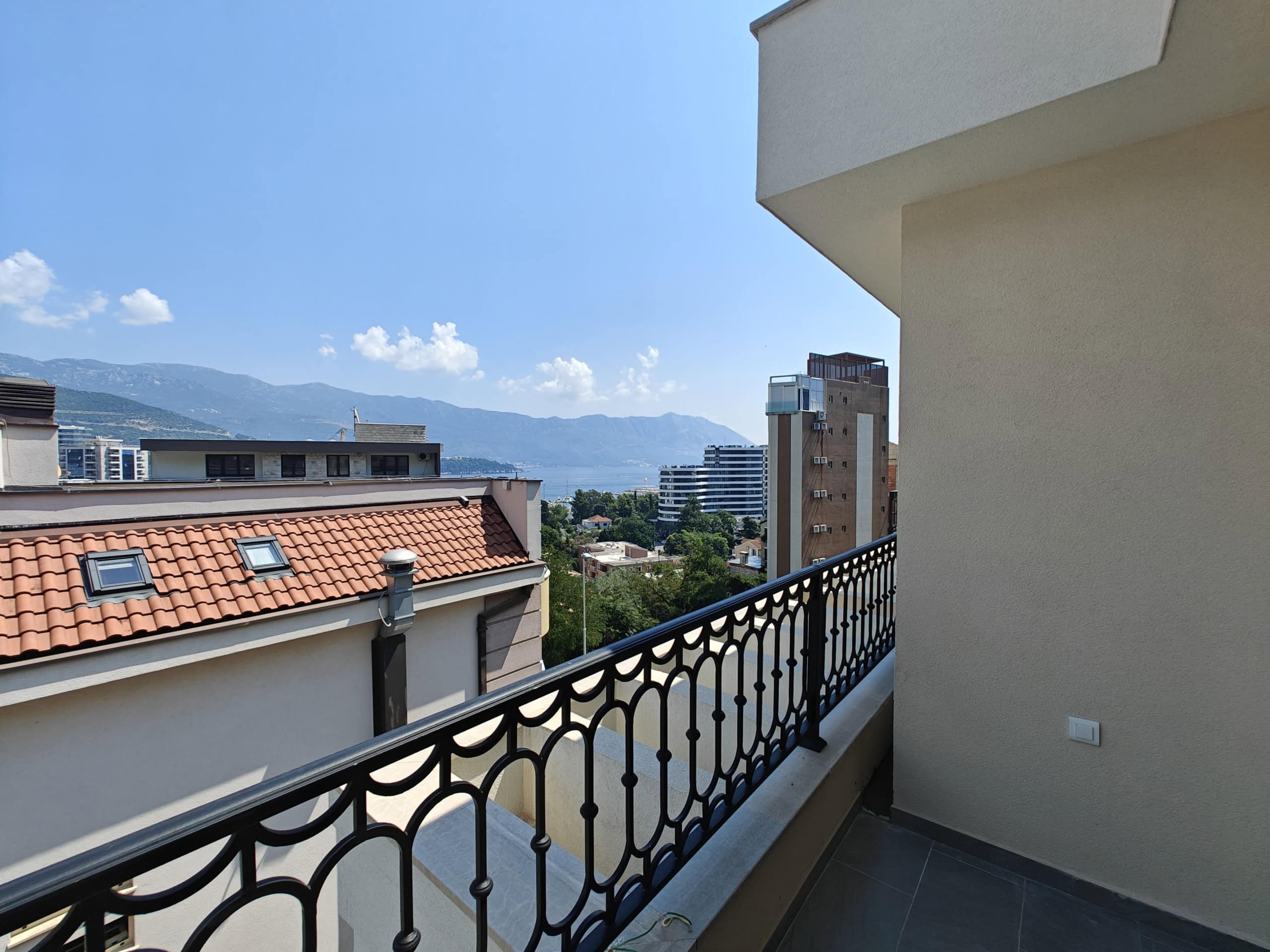 View from an apartment balcony in Budva, Montenegro: panoramic view of the city, mountains, and sea, wrought-iron railing, terracotta roof.