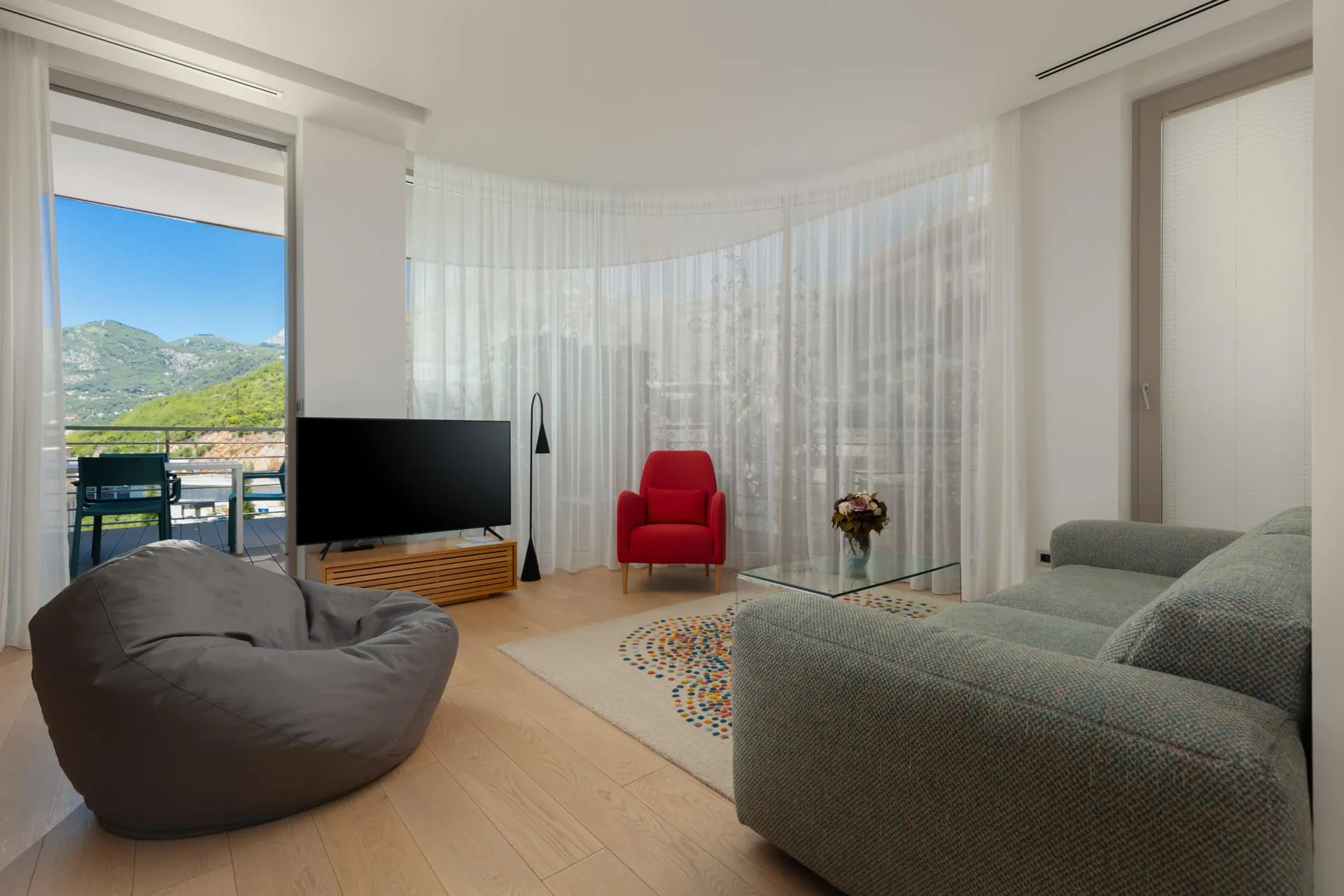 Bright room with a sofa, red armchair, TV, and balcony door overlooking green mountains.