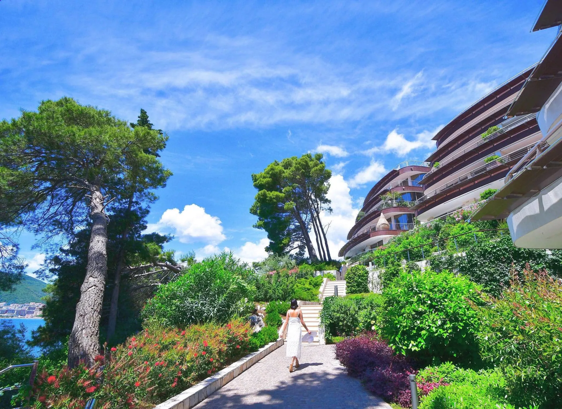 A woman walks along a paved garden path towards a curved modern building with balconies near the sea.