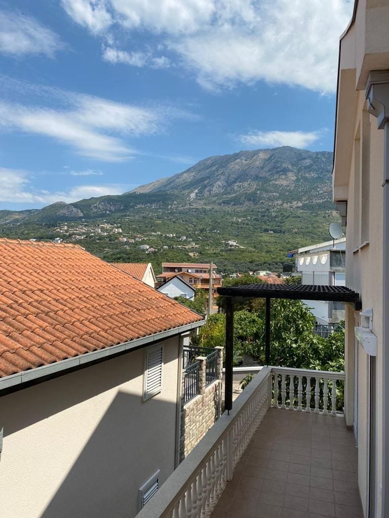 Balcony with white railing and black pergola overlooking a red tiled roof and a green mountain under a blue sky.