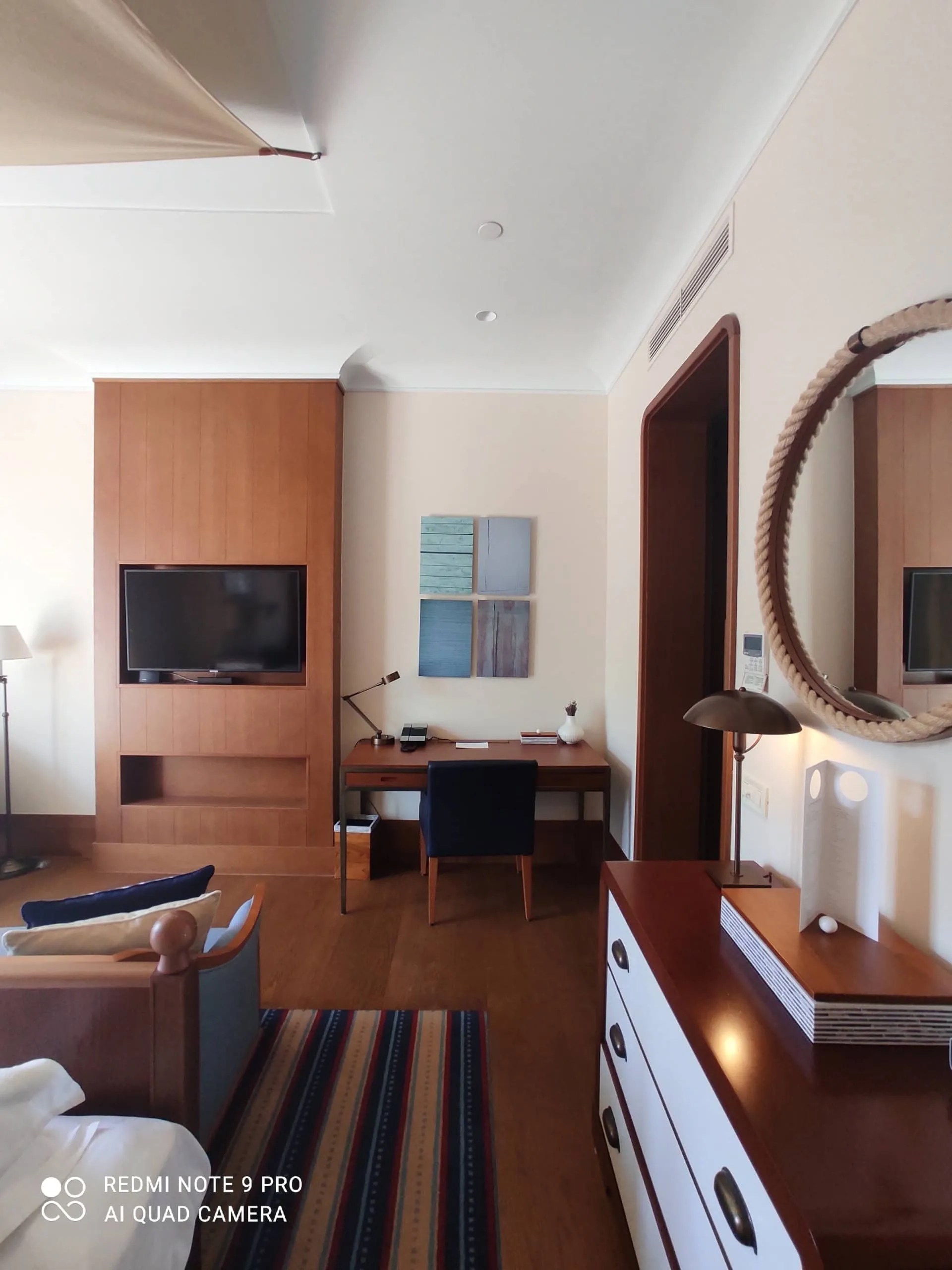 Hotel room featuring wooden furniture, a work desk, wall-mounted TV, and a round rope-framed mirror above a dresser.