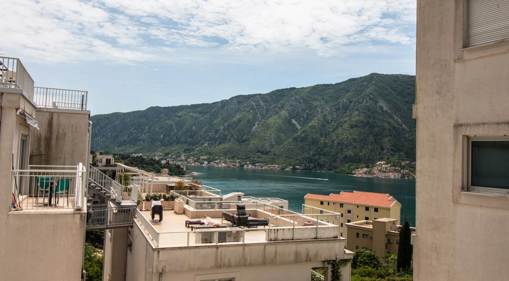 View of a rooftop terrace with furniture, sea, and green mountains. Adjacent buildings visible.