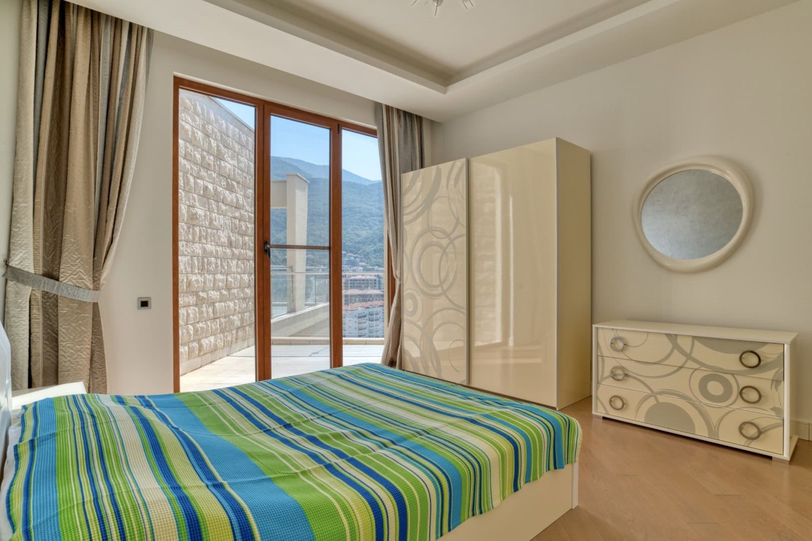 Bedroom in a penthouse in Becici, Montenegro: light-colored furniture, panoramic windows with mountain views, and access to the terrace.