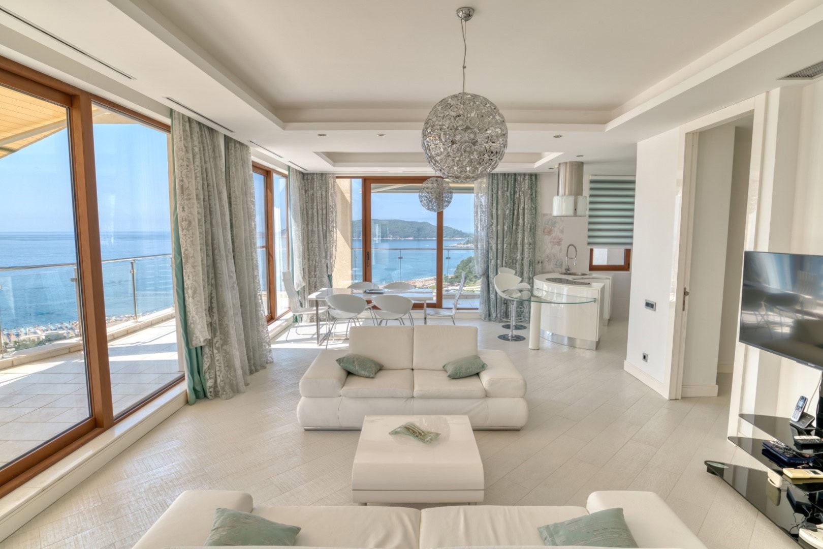 Living room in a penthouse in Becici, Montenegro: panoramic sea view, white furniture, and a modern open-plan kitchen.